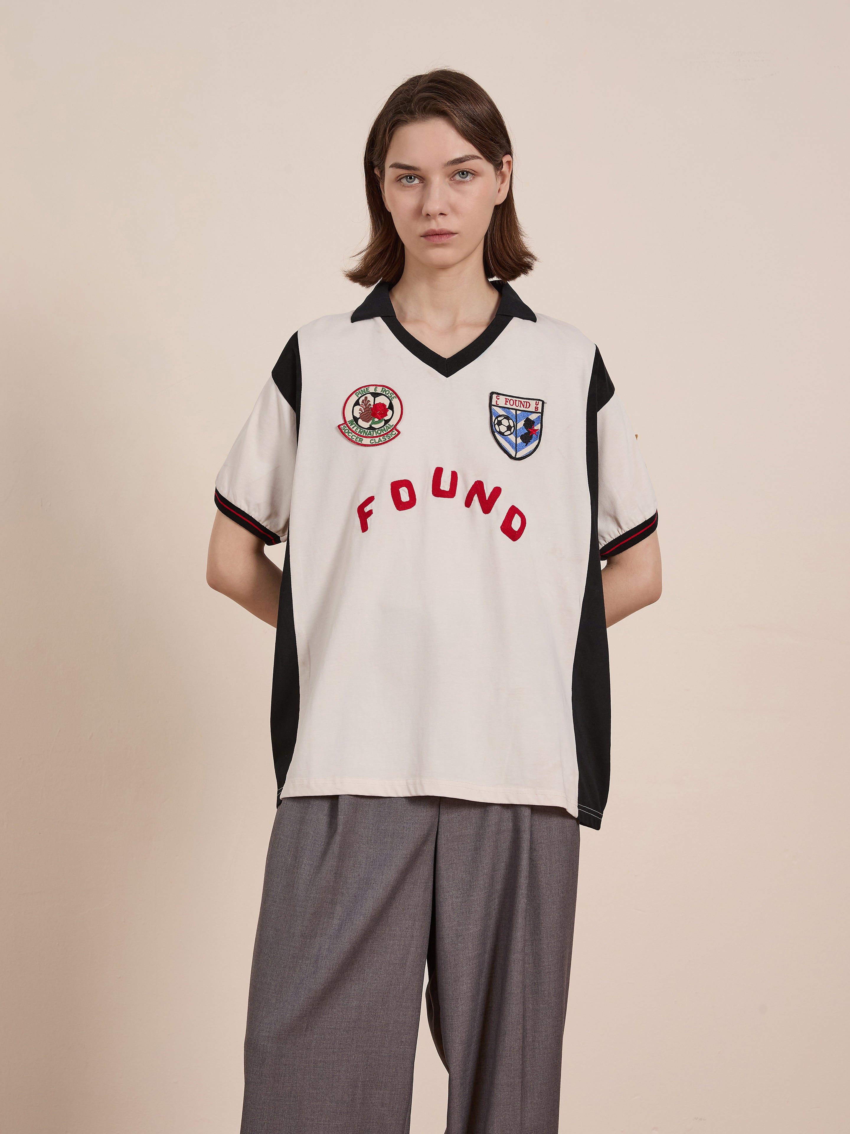 A person with short hair wears a Union Soccer Jersey by FOUND, a white shirt featuring patches and "FOUND" in red, evoking vintage football style. Paired with loose gray pants, they stand against a plain background exuding contemporary edge.