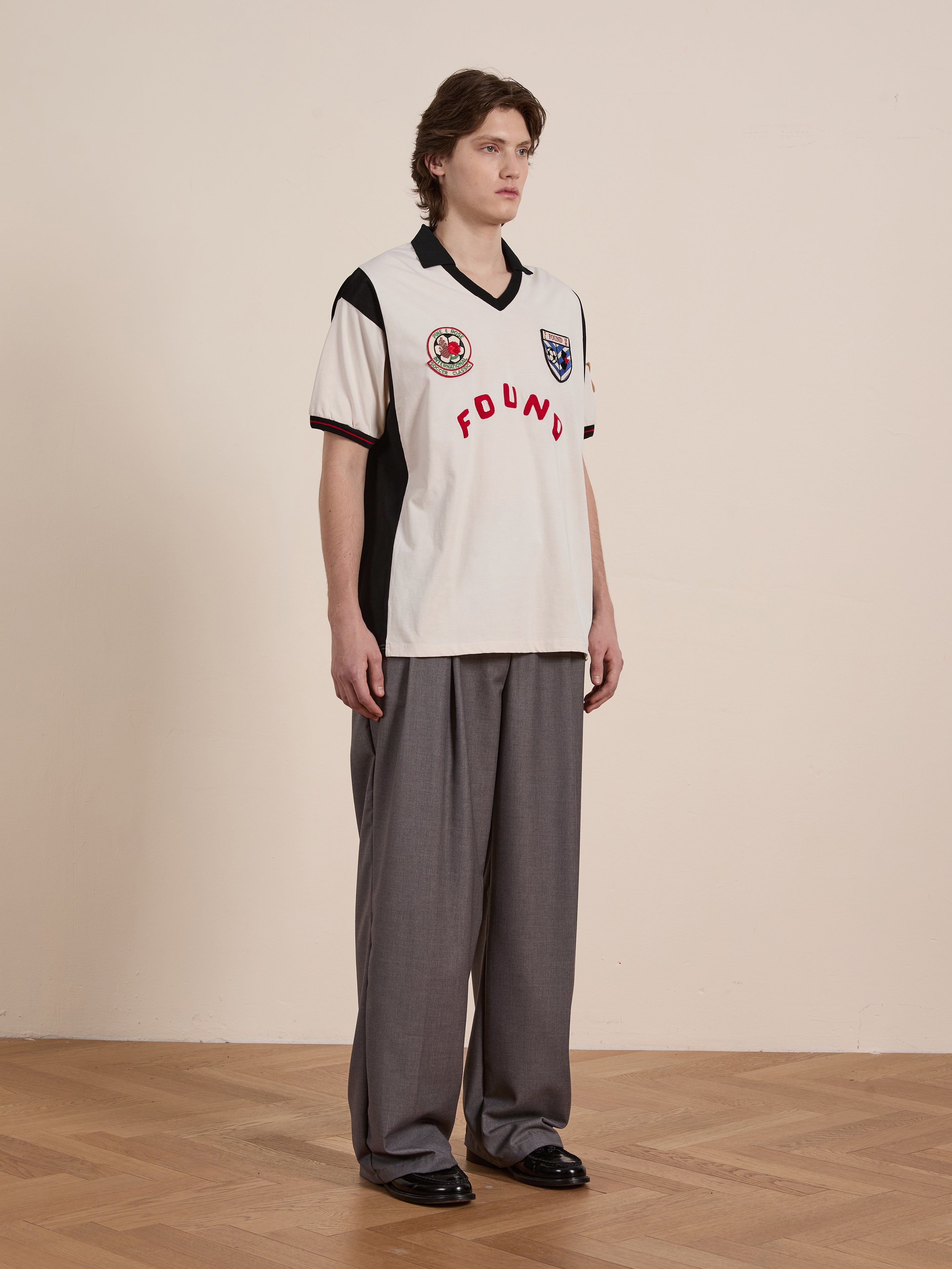 On a wooden floor, a person wears the Union Soccer Jersey by FOUND—a white and black piece paired with gray wide-leg trousers and black shoes—blending contemporary flair with vintage football vibes, facing slightly to the right.