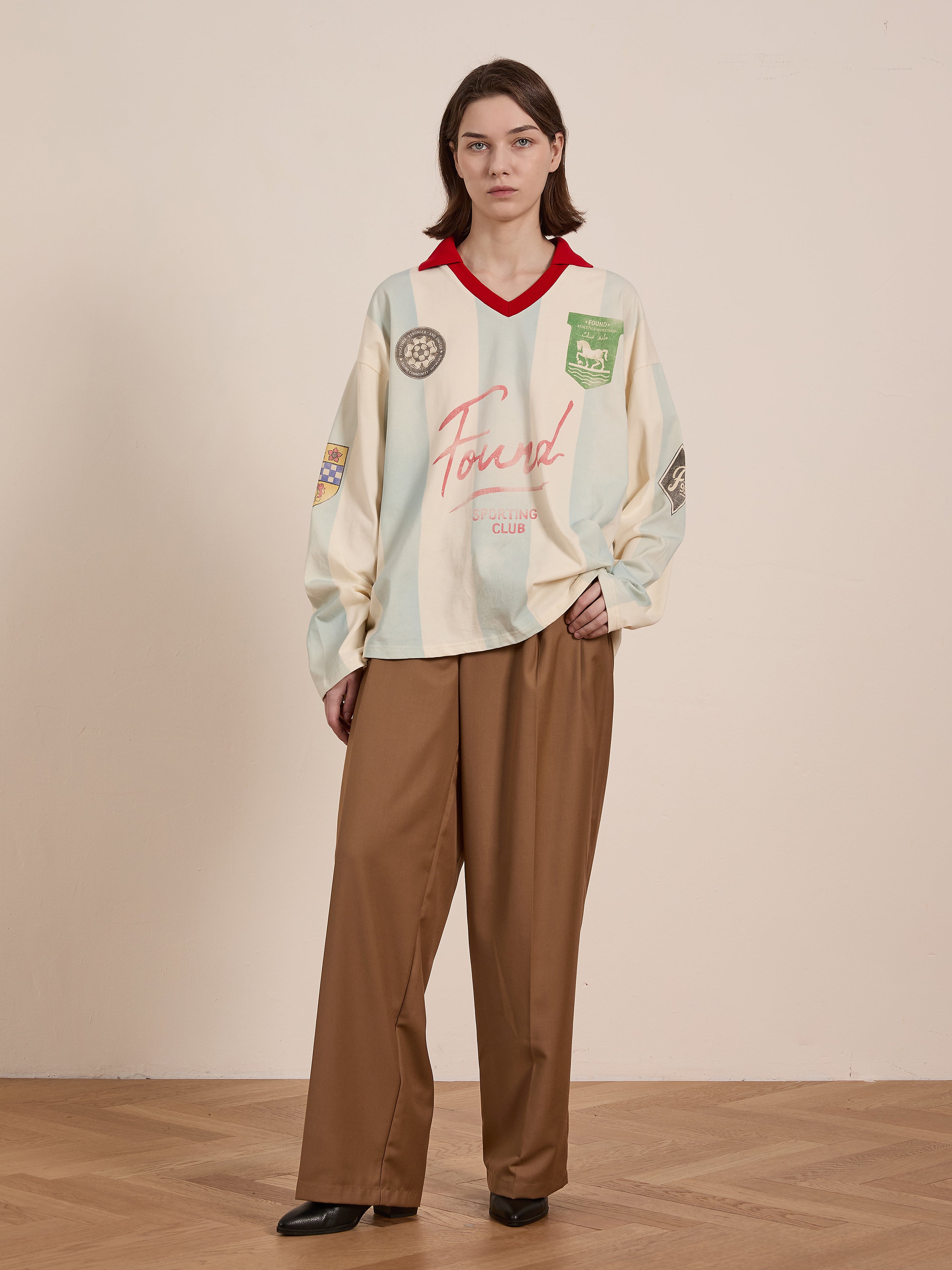 A person indoors displays the FOUND Striped Sport LS Jersey, reminiscent of vintage sportswear, paired with brown pants, on a wooden floor against a plain wall.