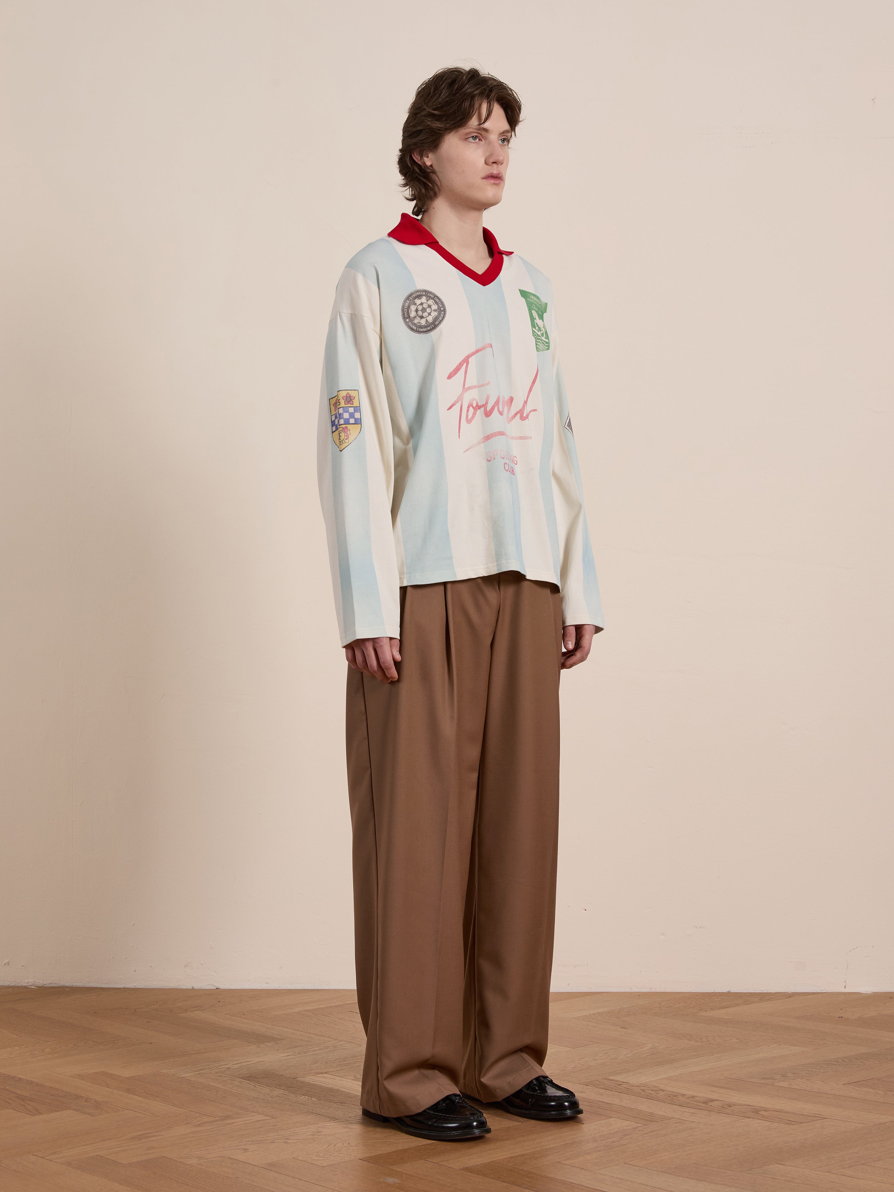 A person stands against a beige wall, wearing the FOUND Striped Sport LS Jersey and brown wide-leg pants, reminiscent of vintage sportswear, on a wooden floor.