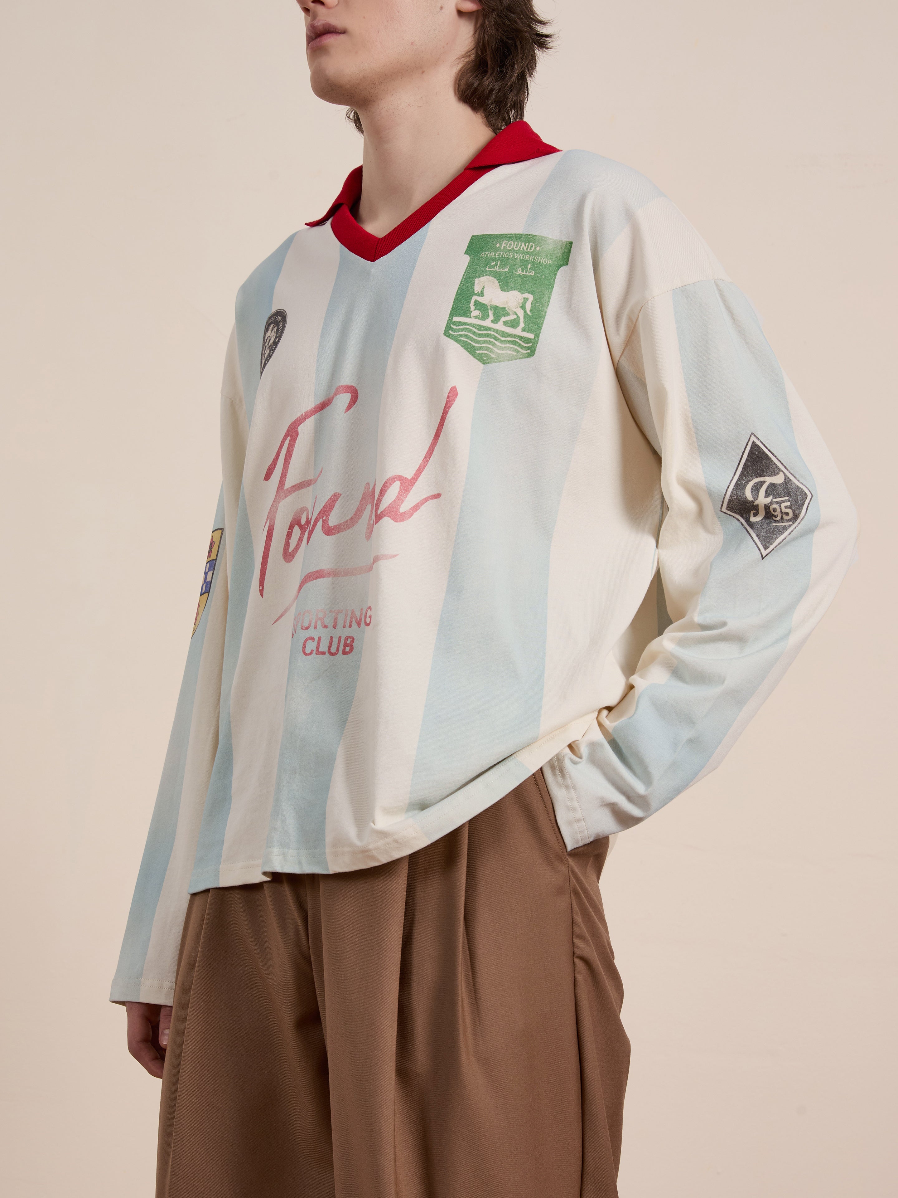 The person sports a vintage look with FOUND's Striped Sport LS Jersey, featuring "Ford Sporting Club" text and various patches, stylishly paired with brown pants.