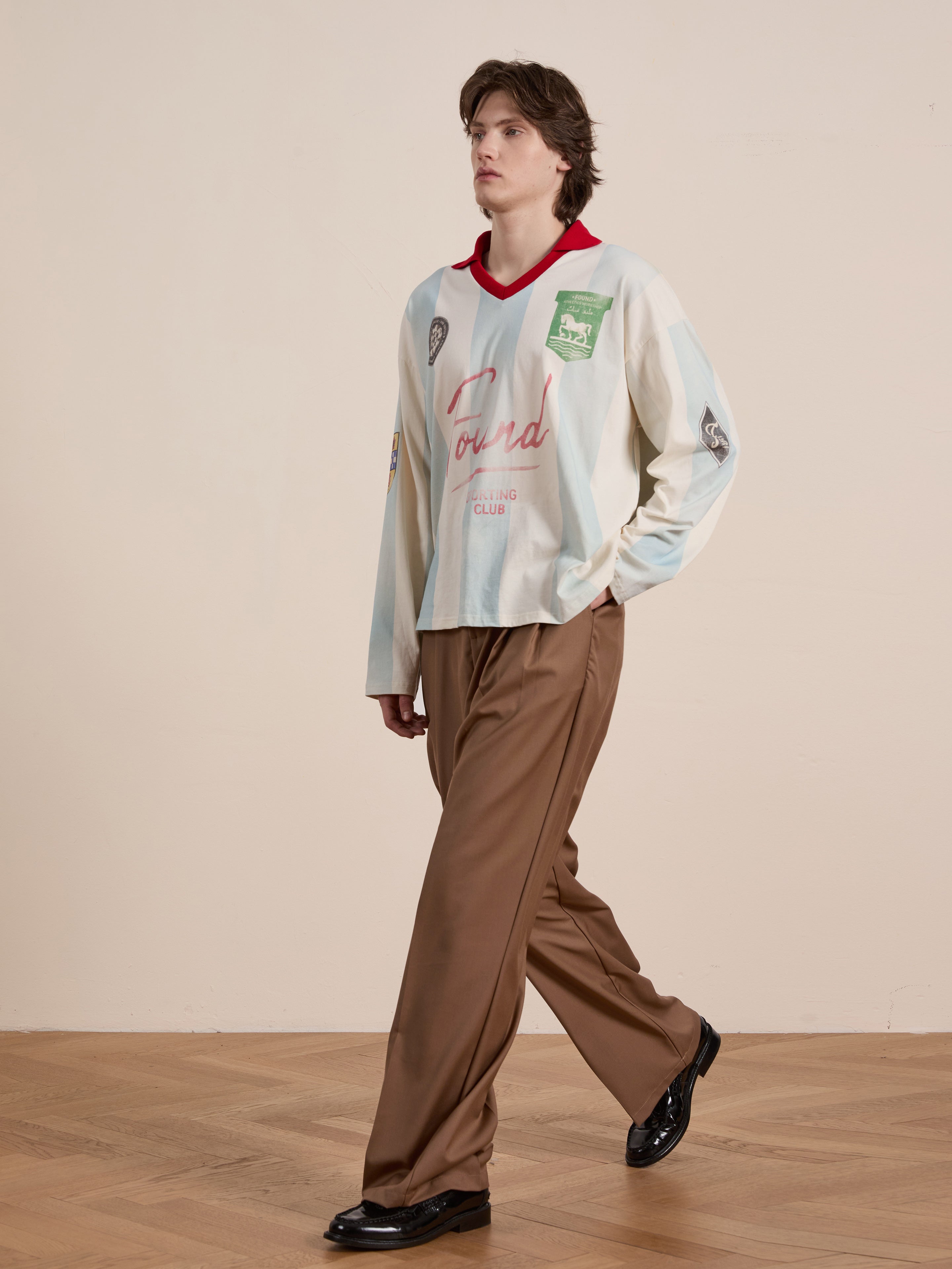 A person wearing the FOUND Striped Sport LS Jersey, with cream and red vintage sportswear vibes, paired with brown wide-leg pants and black shoes, walks on a wooden floor.