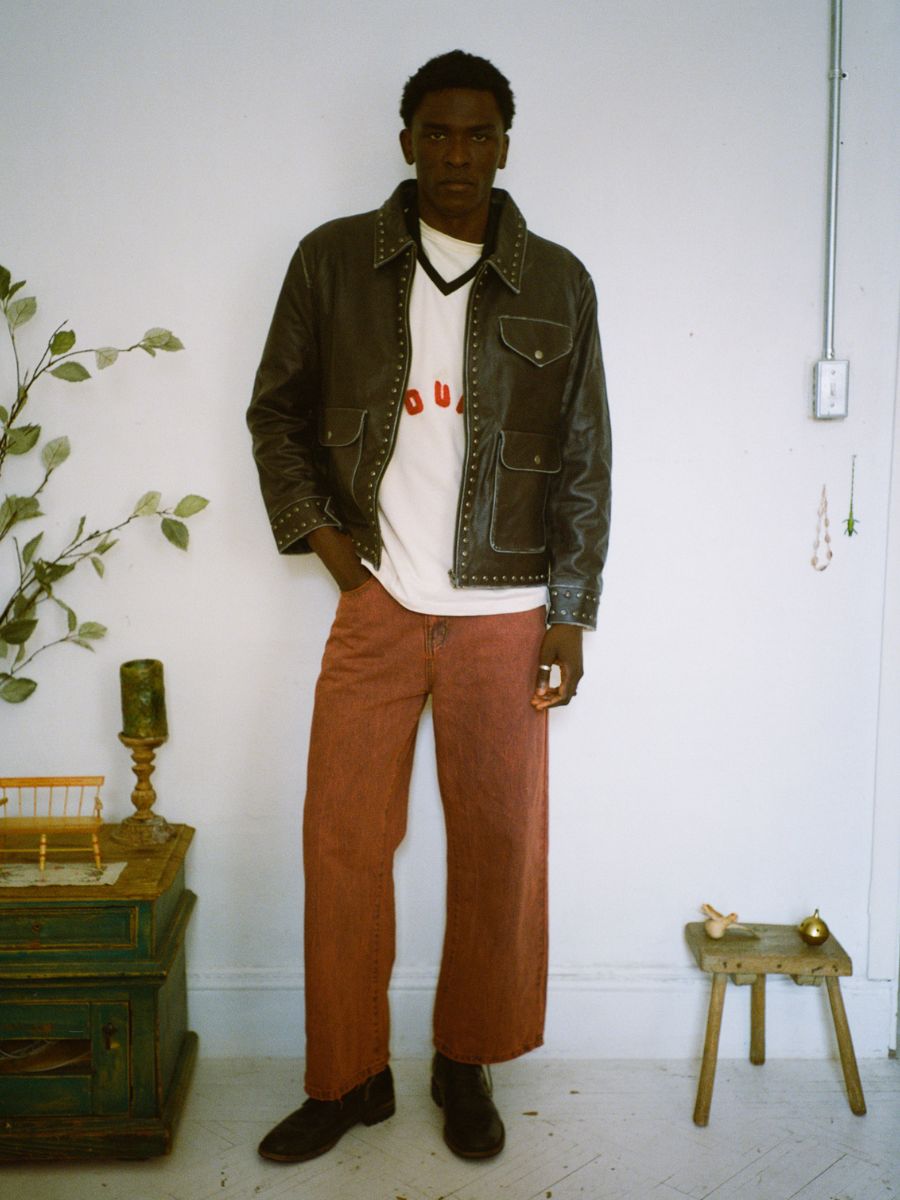 A person stands in a room, wearing FOUND's Studded Leather Pocket Jacket over a white shirt and red pants. The unisex relaxed fit enhances the scene flawlessly, complemented by a small wooden stool and decorative items contributing to the stylish ambiance.