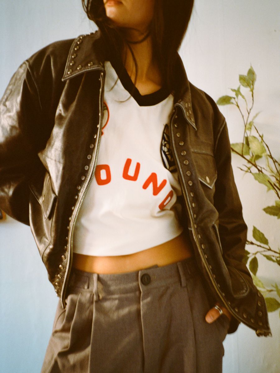 Wearing FOUND's Studded Leather Pocket Jacket, this outfit includes a white cropped shirt with bold red letters and unisex relaxed-fit gray pleated trousers.