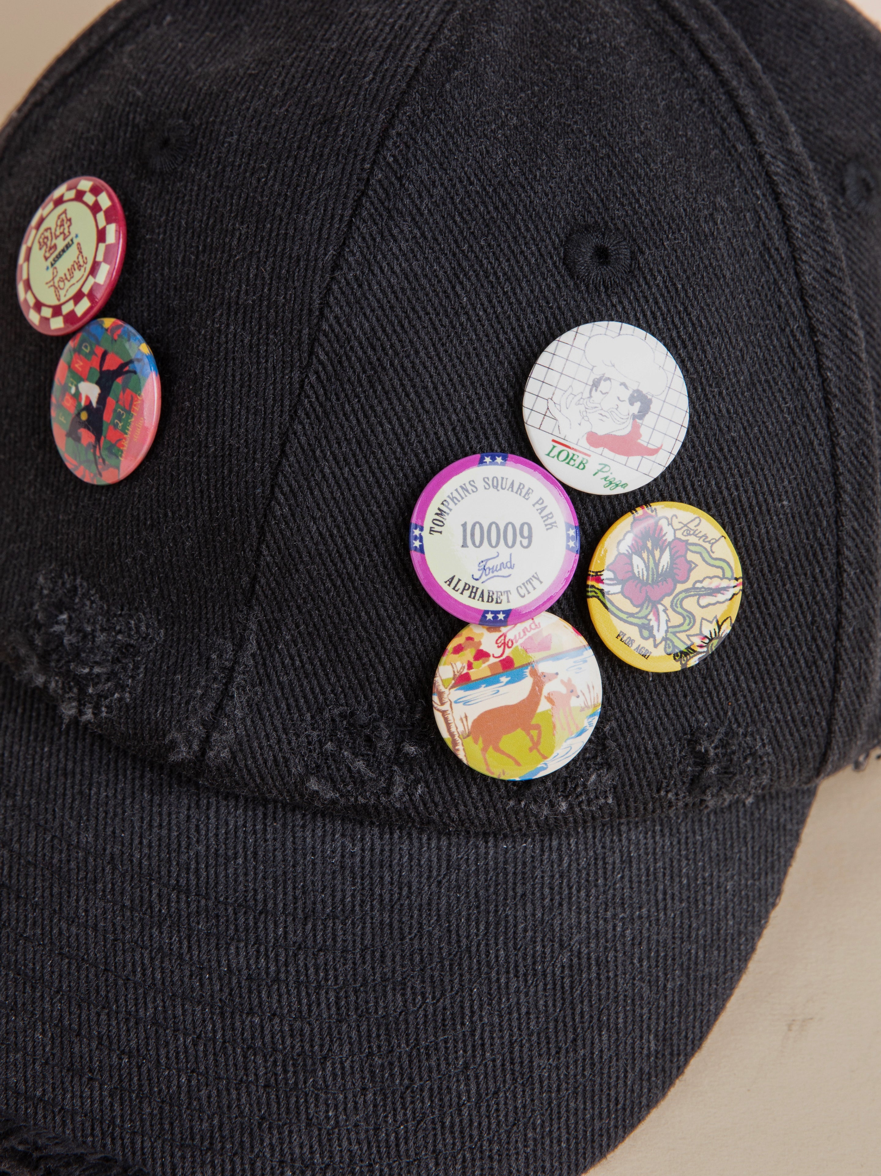 The Distressed Pin Cap by FOUND is a black cotton cap embellished with various colorful, vintage-inspired pins that feature designs and text such as "Alphabet City" and other illustrations.