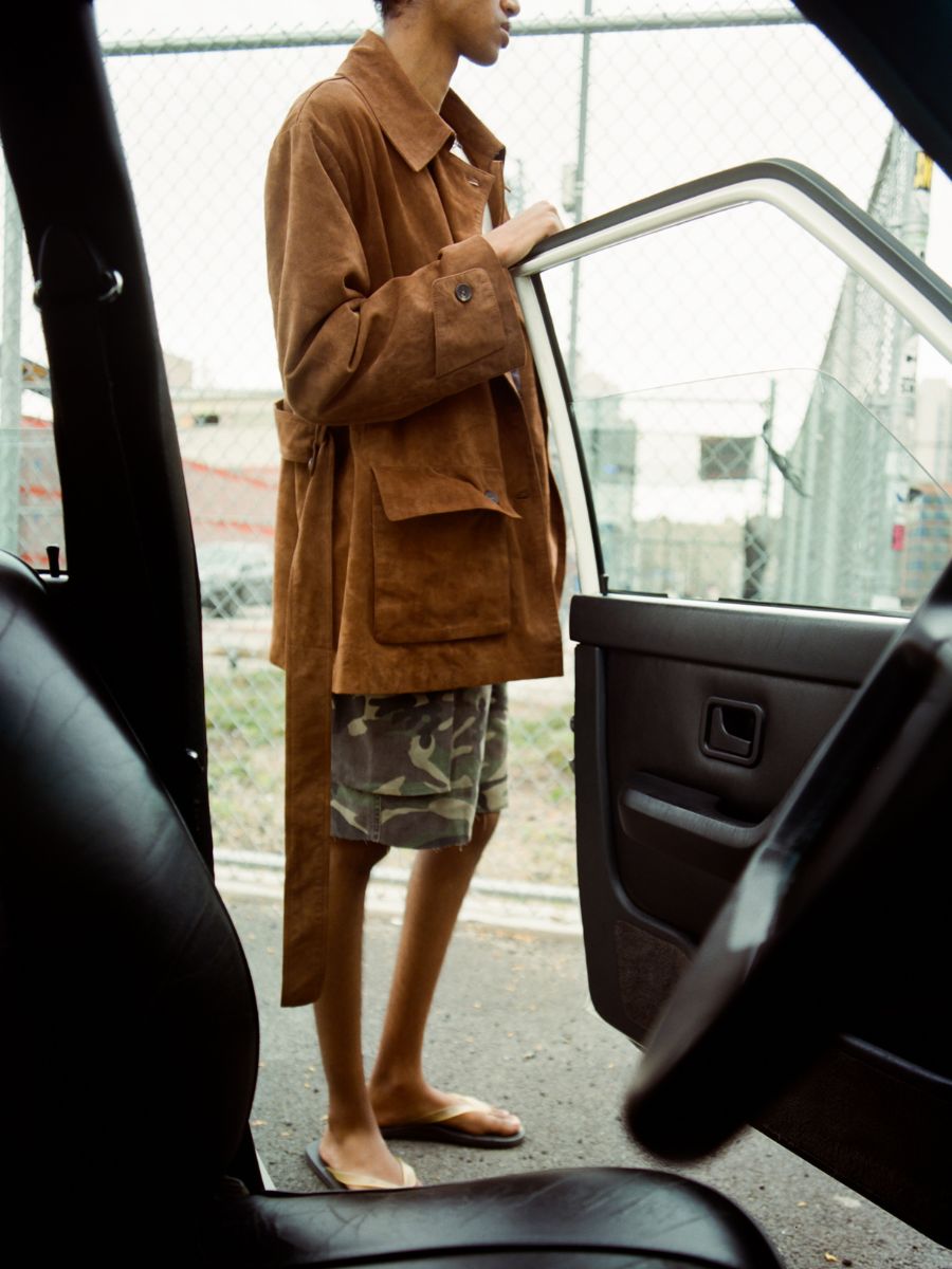 Wearing the FOUND Double Breasted Suede Leather Jacket, camouflage shorts, and flip-flops, a person stands outside an open car door near a chain-link fence.