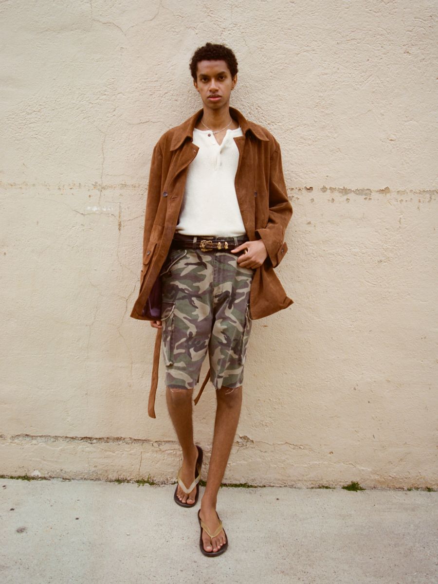 A person stands against a beige wall, wearing the FOUND Double Breasted Suede Leather Jacket in brown, paired with a white shirt, camouflage shorts, a belt, and flip-flops.