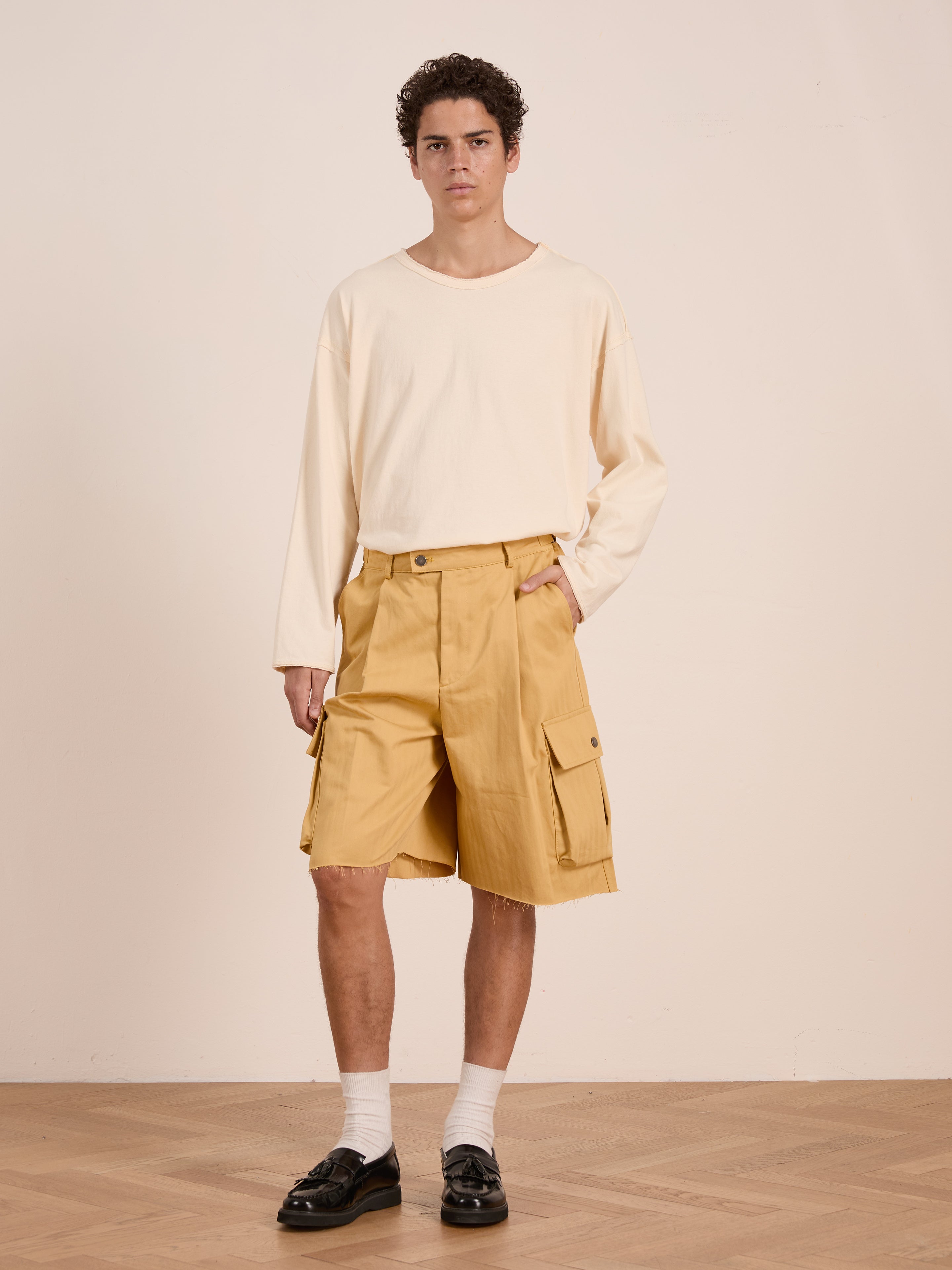 A person stands indoors by a plain light wall, wearing a cream long-sleeve shirt, FOUND Sunlight Twill Cargo Shorts in yellow, white socks, and black loafers.