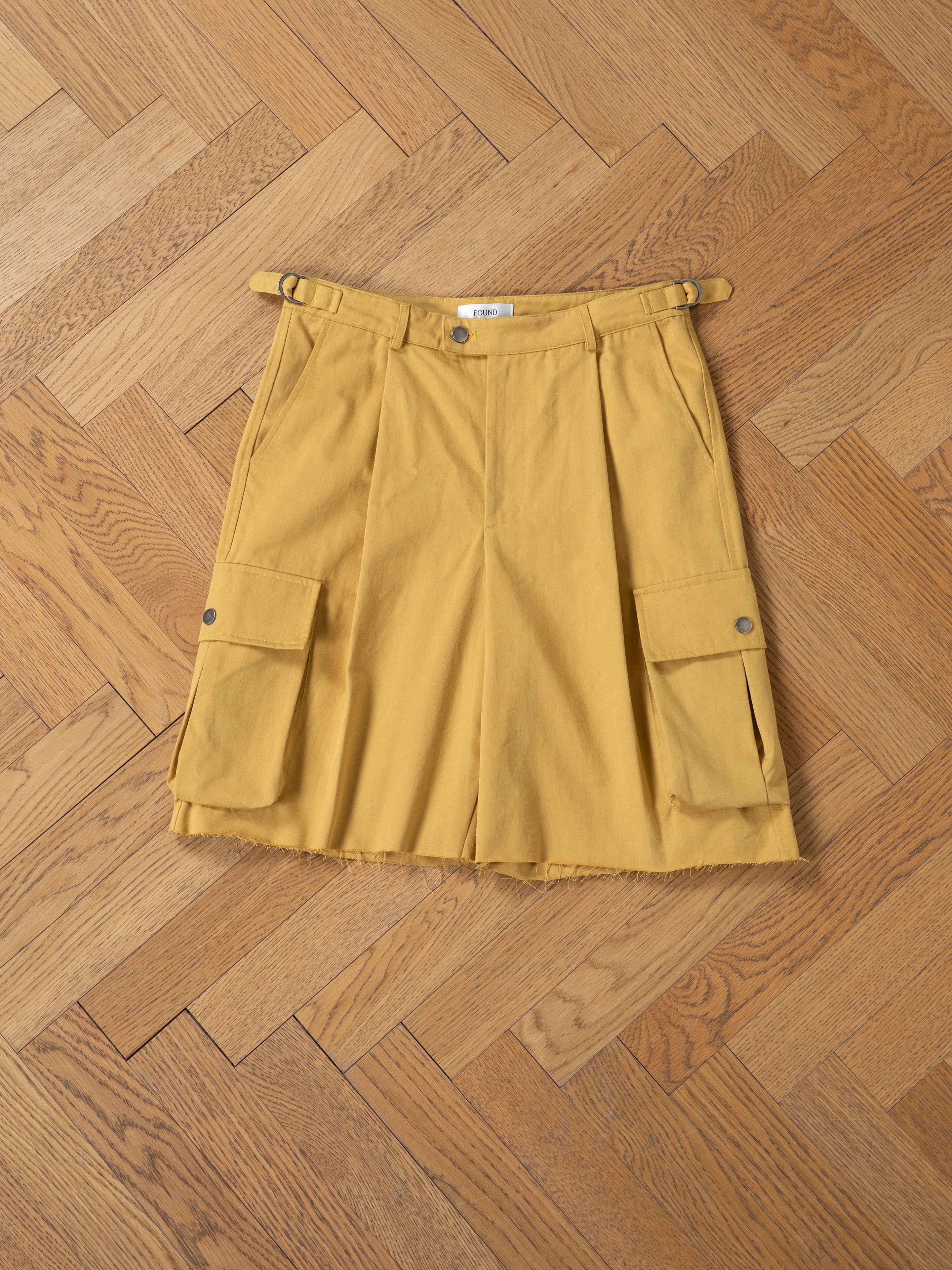The FOUND Sunlight Twill Cargo Shorts are unisex, crafted from herringbone twill in mustard yellow, featuring front pleats and two large side pockets, shown laid flat on a wooden herringbone floor.