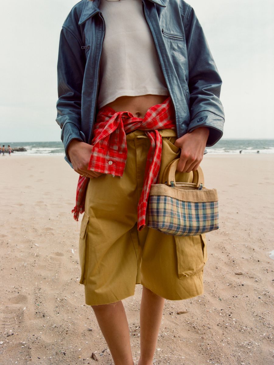 A person stands on a beach in a blue jacket, white crop top, red plaid shirt tied at the waist, FOUND Sunlight Twill Cargo Shorts, and holding a small plaid handbag for a stylish unisex look.