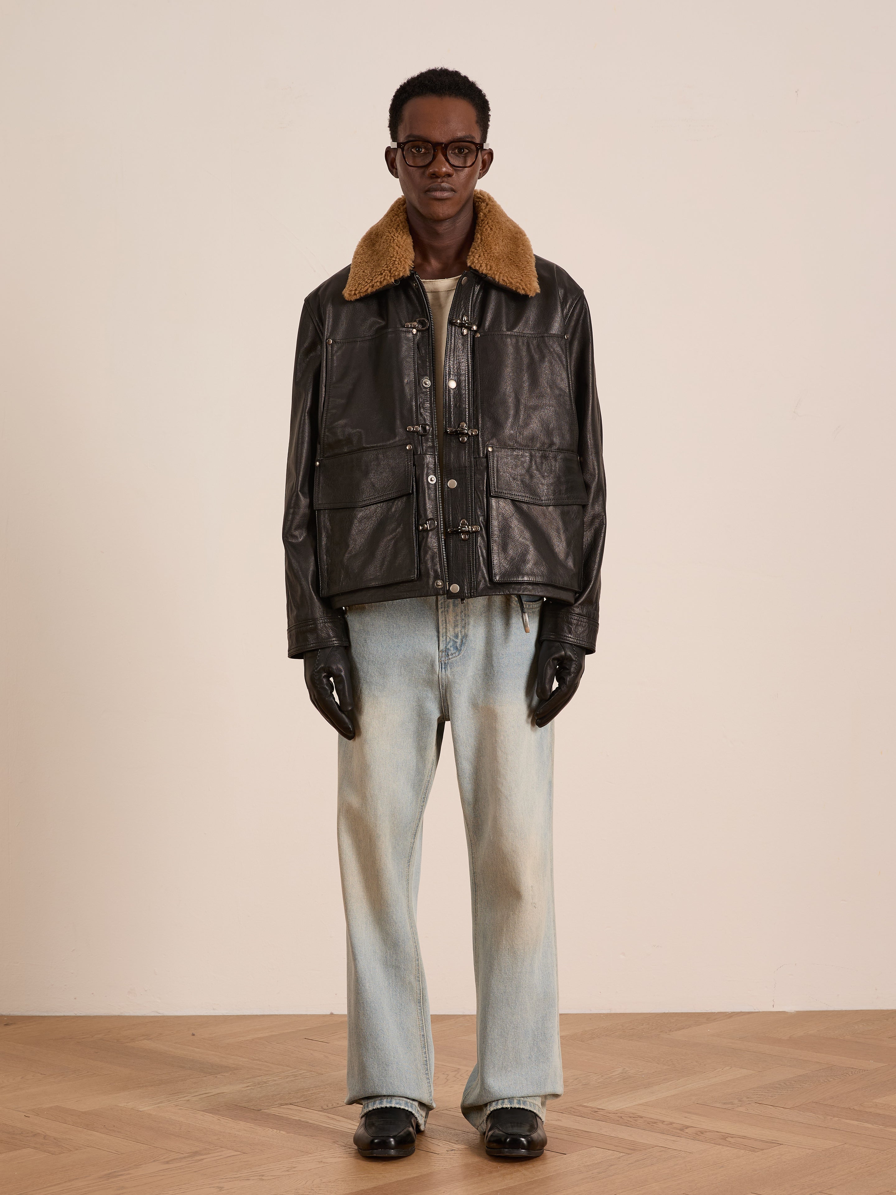 A person stands indoors against a plain background, wearing the FOUND Sherpa Collar Leather Utility Jacket, light washed jeans, black gloves, black shoes, and glasses.