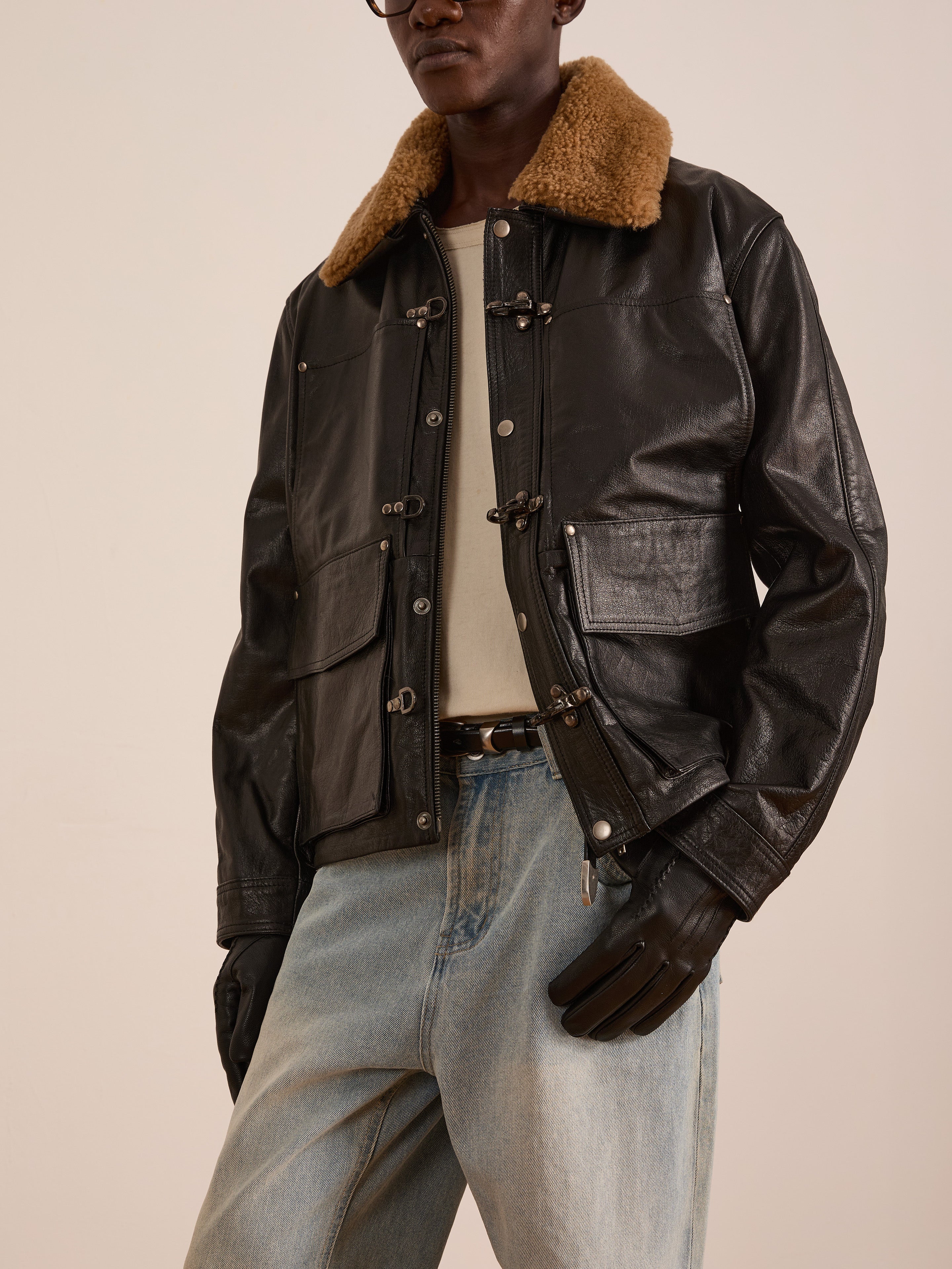 A person wearing the FOUND Sherpa Collar Leather Utility Jacket—a genuine leather piece with a brown sherpa collar—over a light shirt, jeans, and black gloves stands against a plain background.