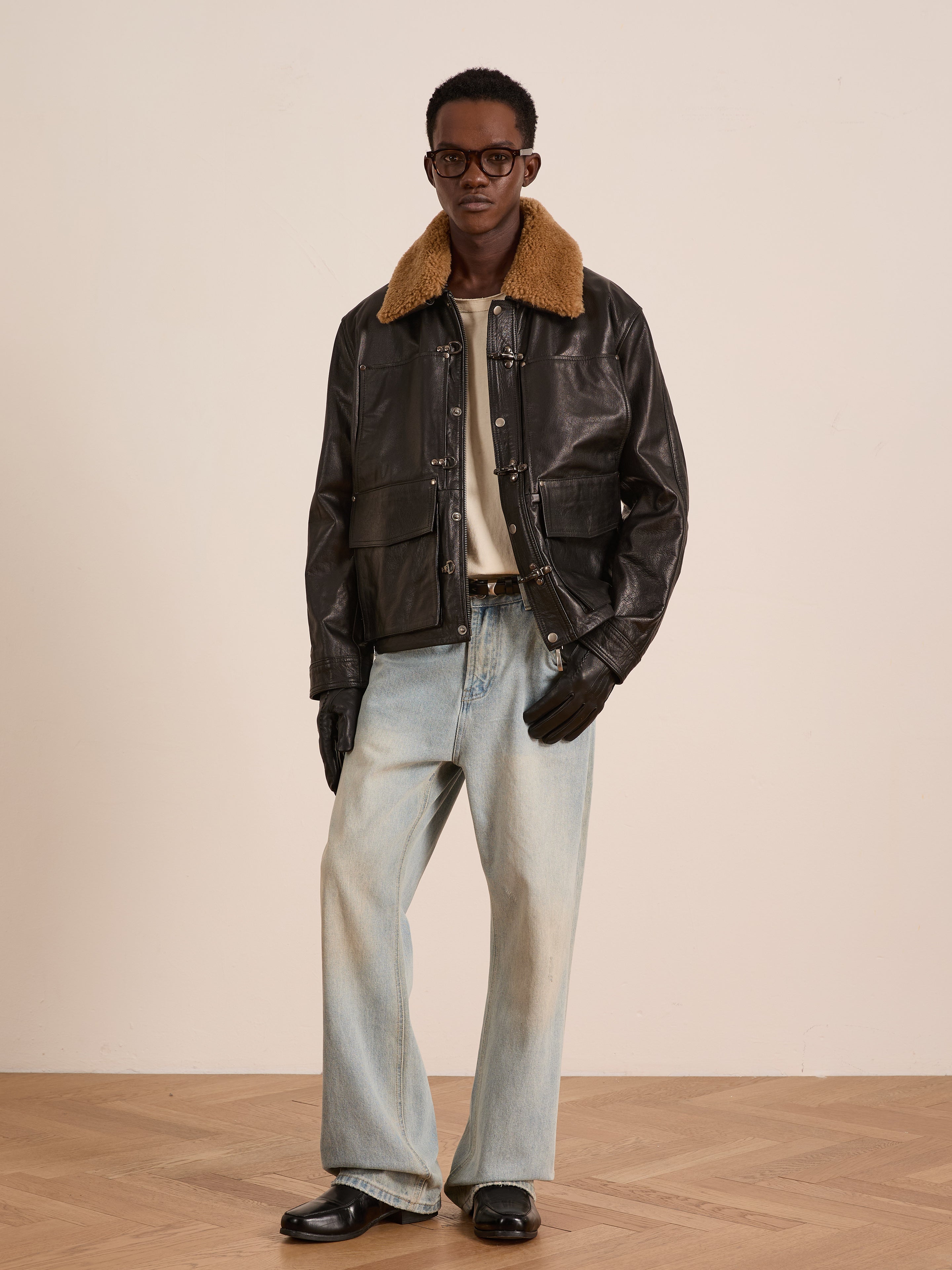 A man stands indoors wearing a FOUND Sherpa Collar Leather Utility Jacket, brown shearling collar, light blue jeans, black gloves, black shoes, white shirt, and round glasses.