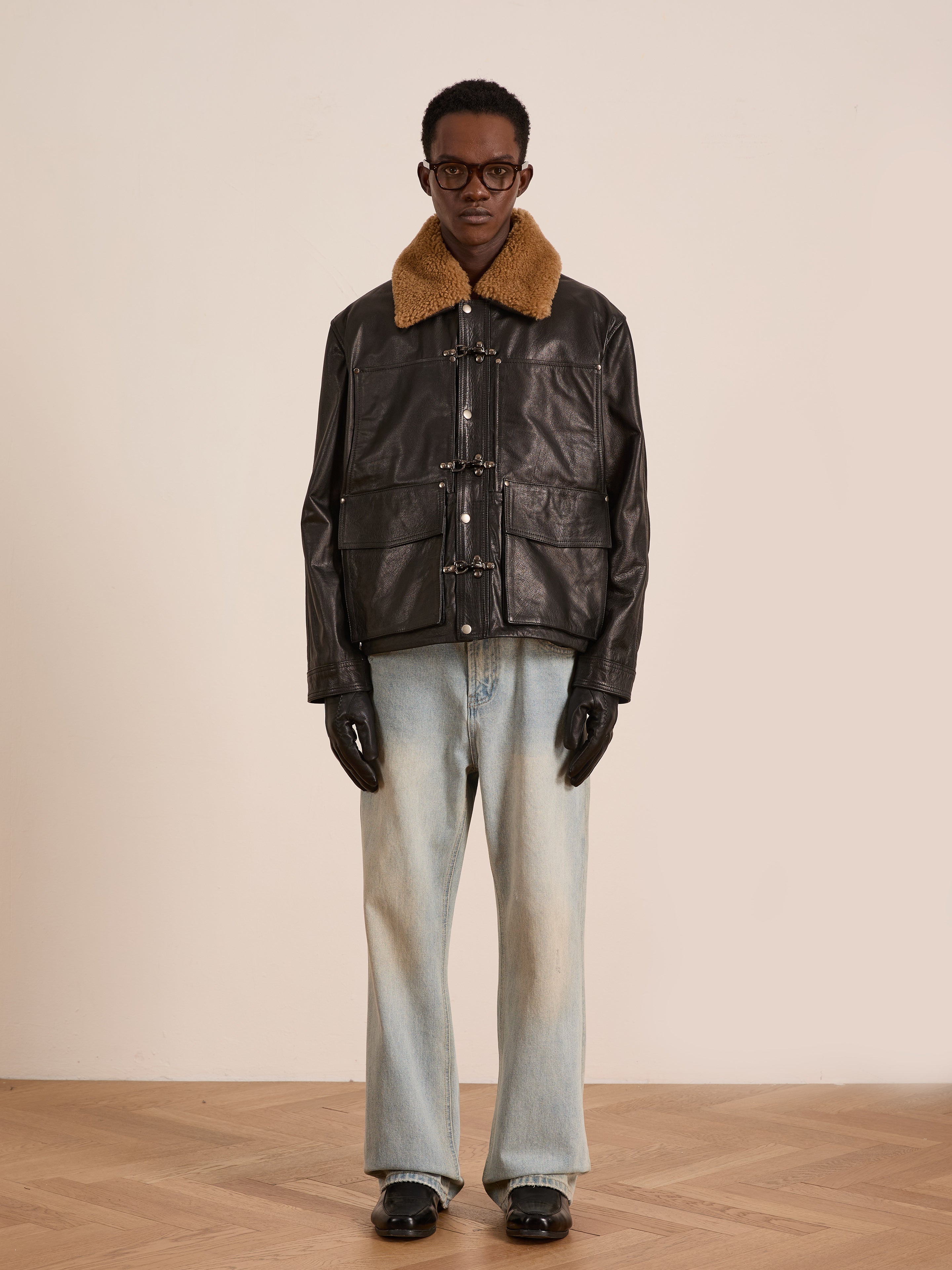 A person wearing glasses, a FOUND Sherpa Collar Leather Utility Jacket, black gloves, light blue jeans, and black shoes stands against a plain background.