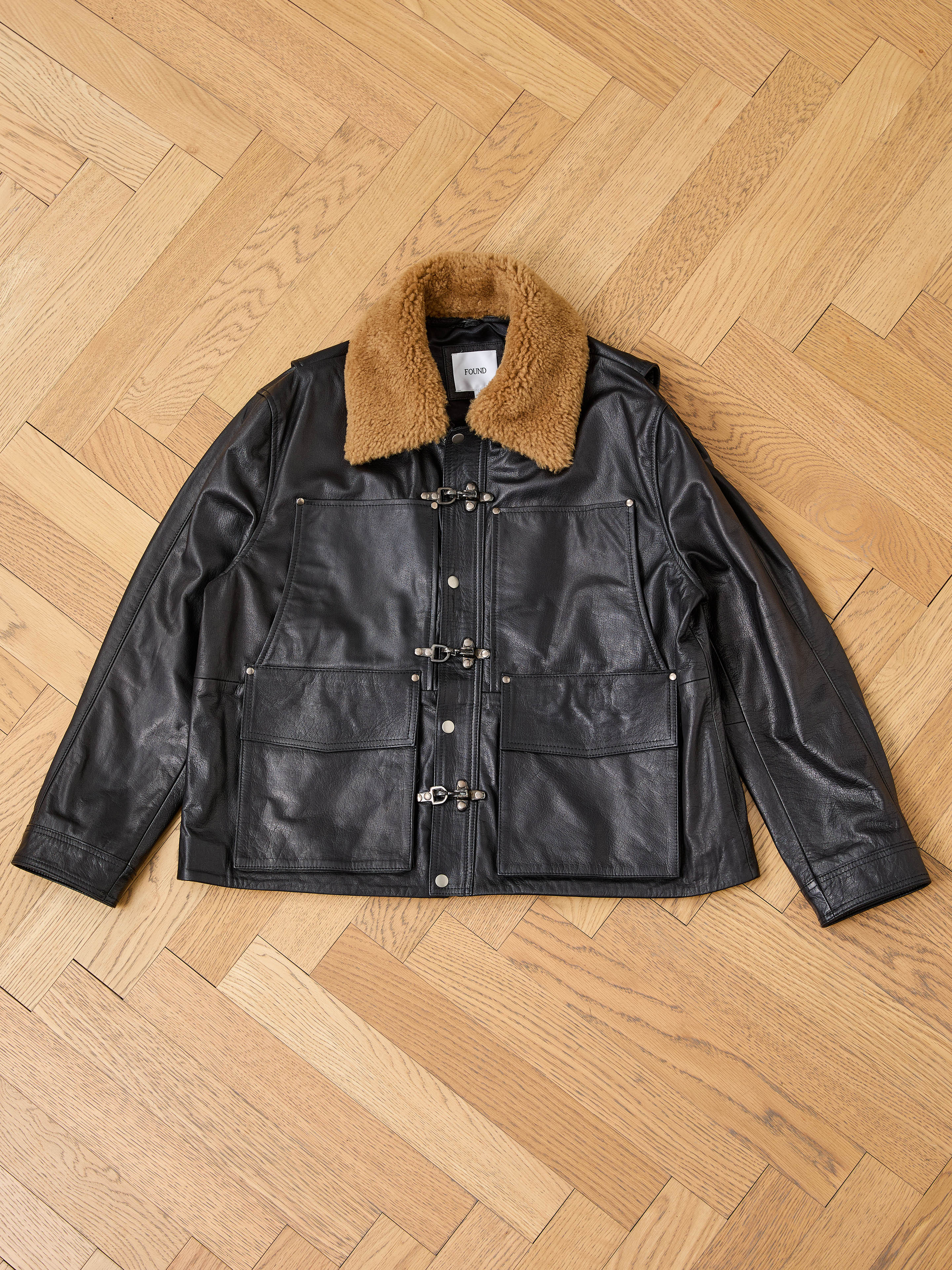 FOUND Sherpa Collar Leather Utility Jacket in black genuine leather with a brown shearling collar, front toggle closures, and four large pockets, shown laid flat on a wooden parquet floor.