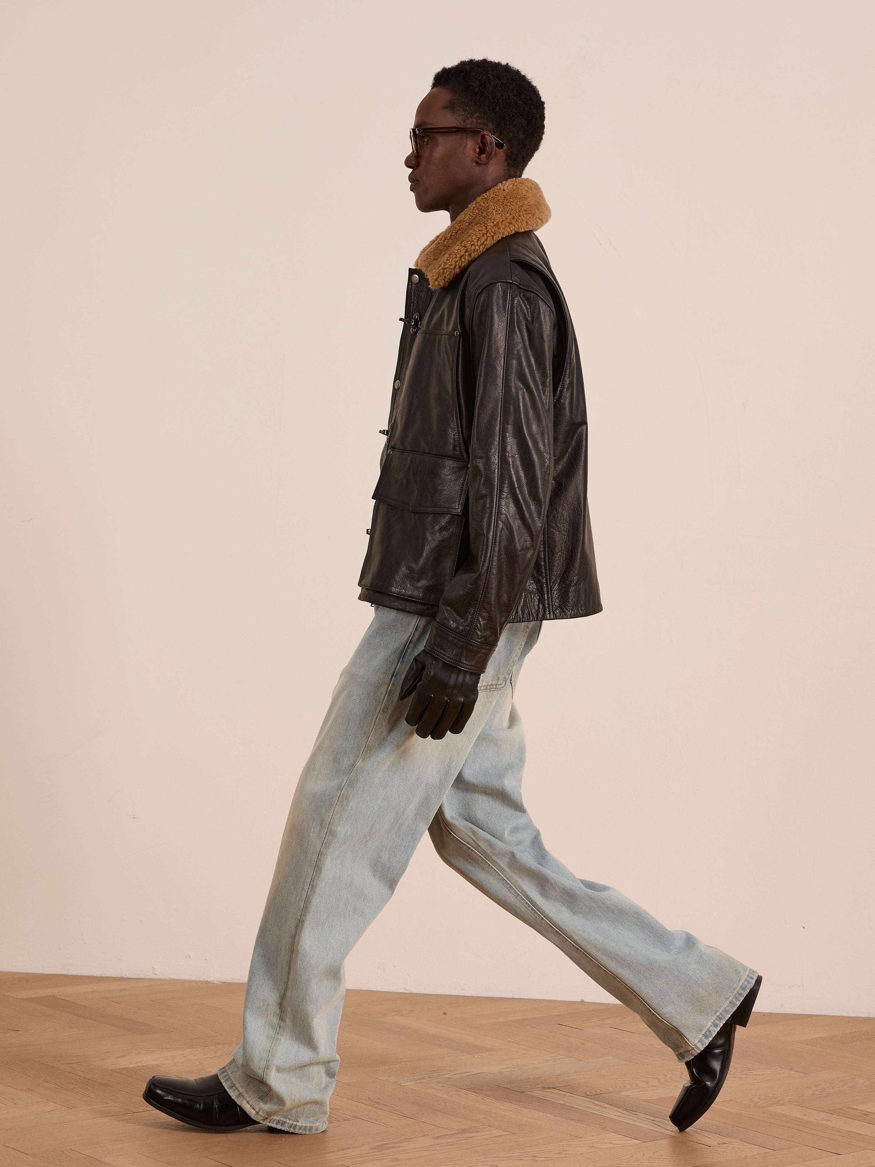 A person wearing the FOUND Sherpa Collar Leather Utility Jacket, light blue jeans, black gloves, and black shoes walks indoors on a wood floor.