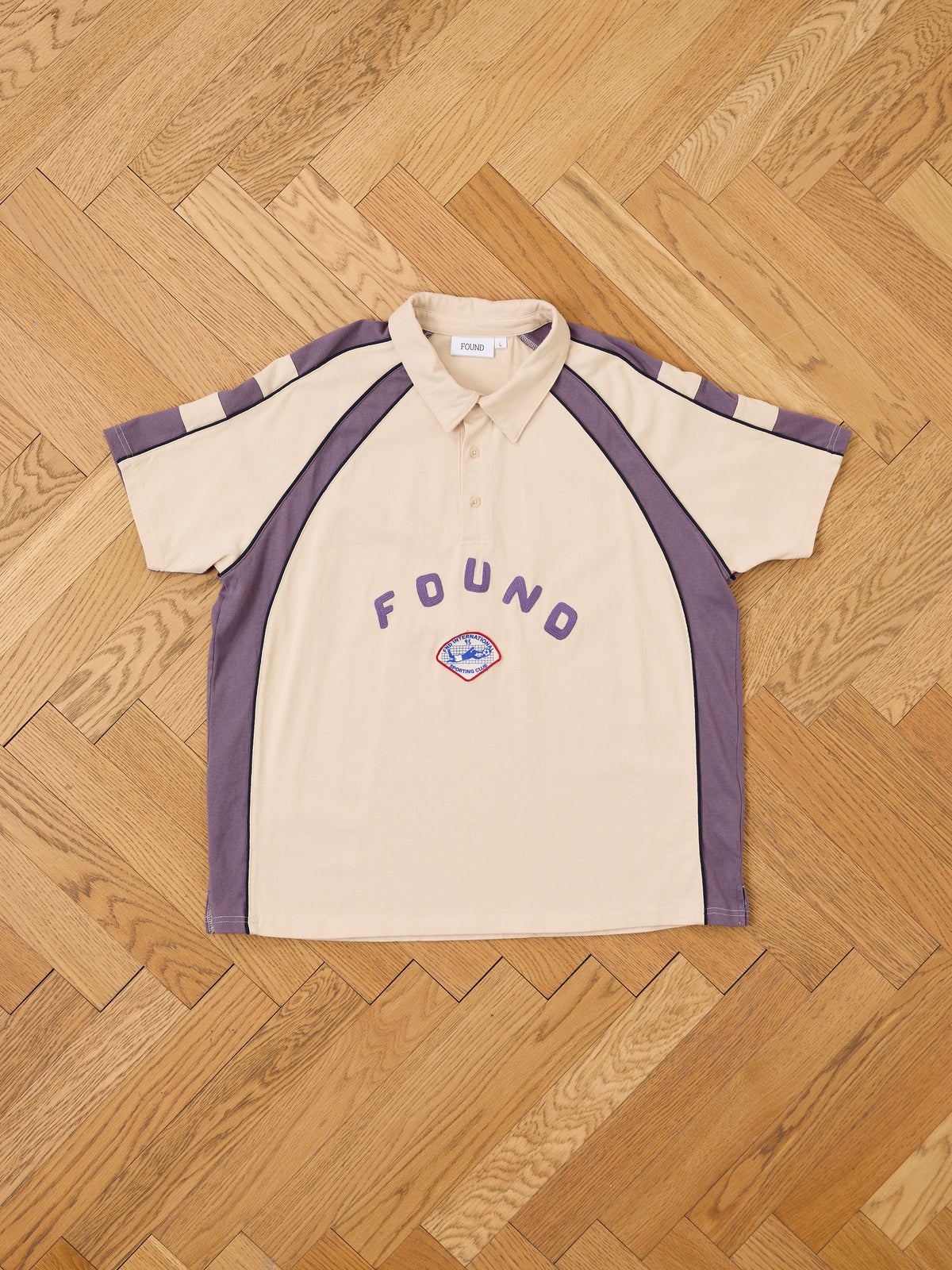 The Short Sleeve Rugby Polo by FOUND in beige and purple, unisex relaxed fit with “FOUND” text and a small embroidered patch, is laid flat on herringbone wood flooring.
