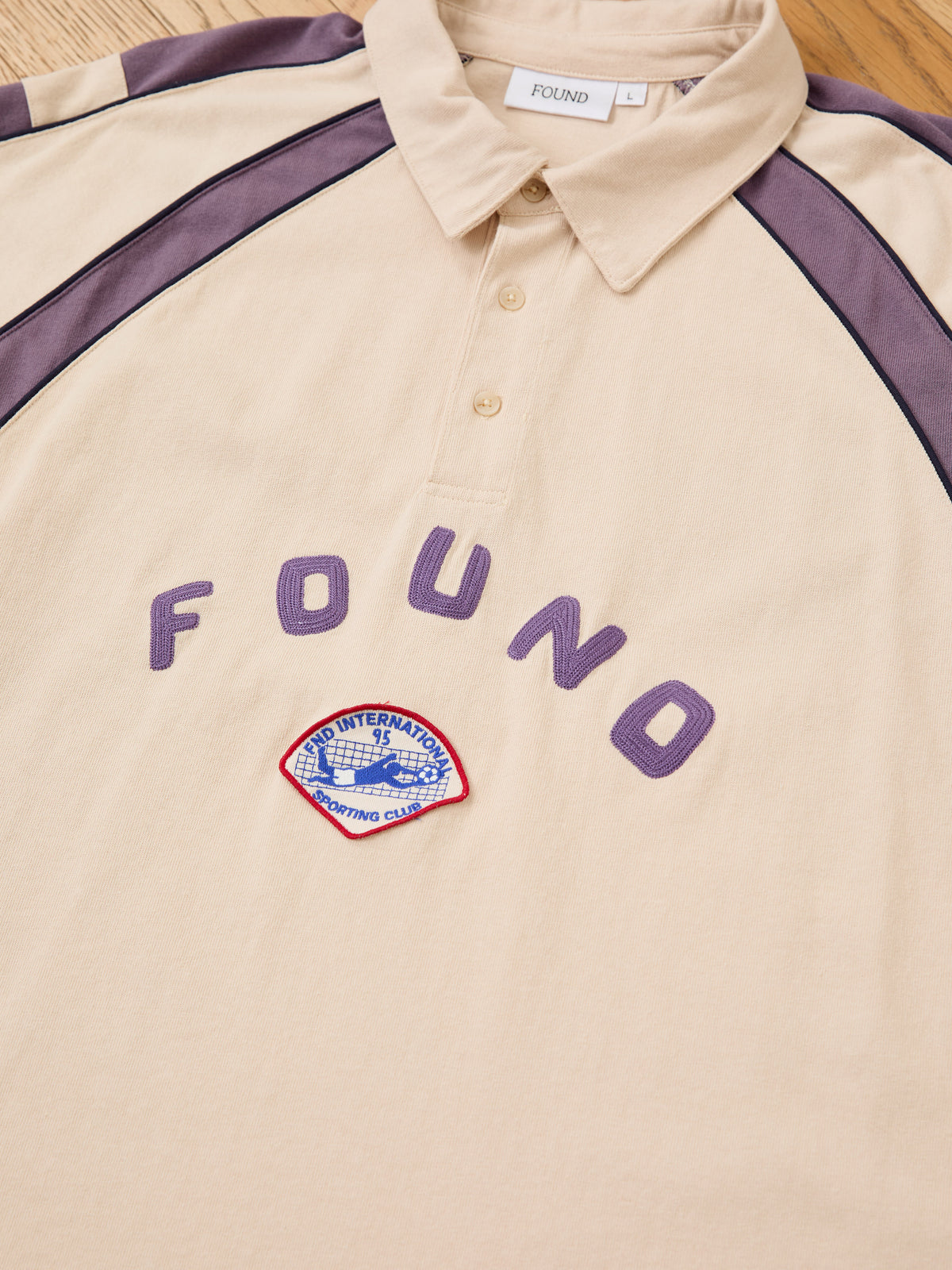Short Sleeve Rugby Polo by FOUND: beige, purple striped shoulders, “FOUND” text, vintage look, “INTERNATIONAL SPORTING CLUB” patch; unisex relaxed fit.