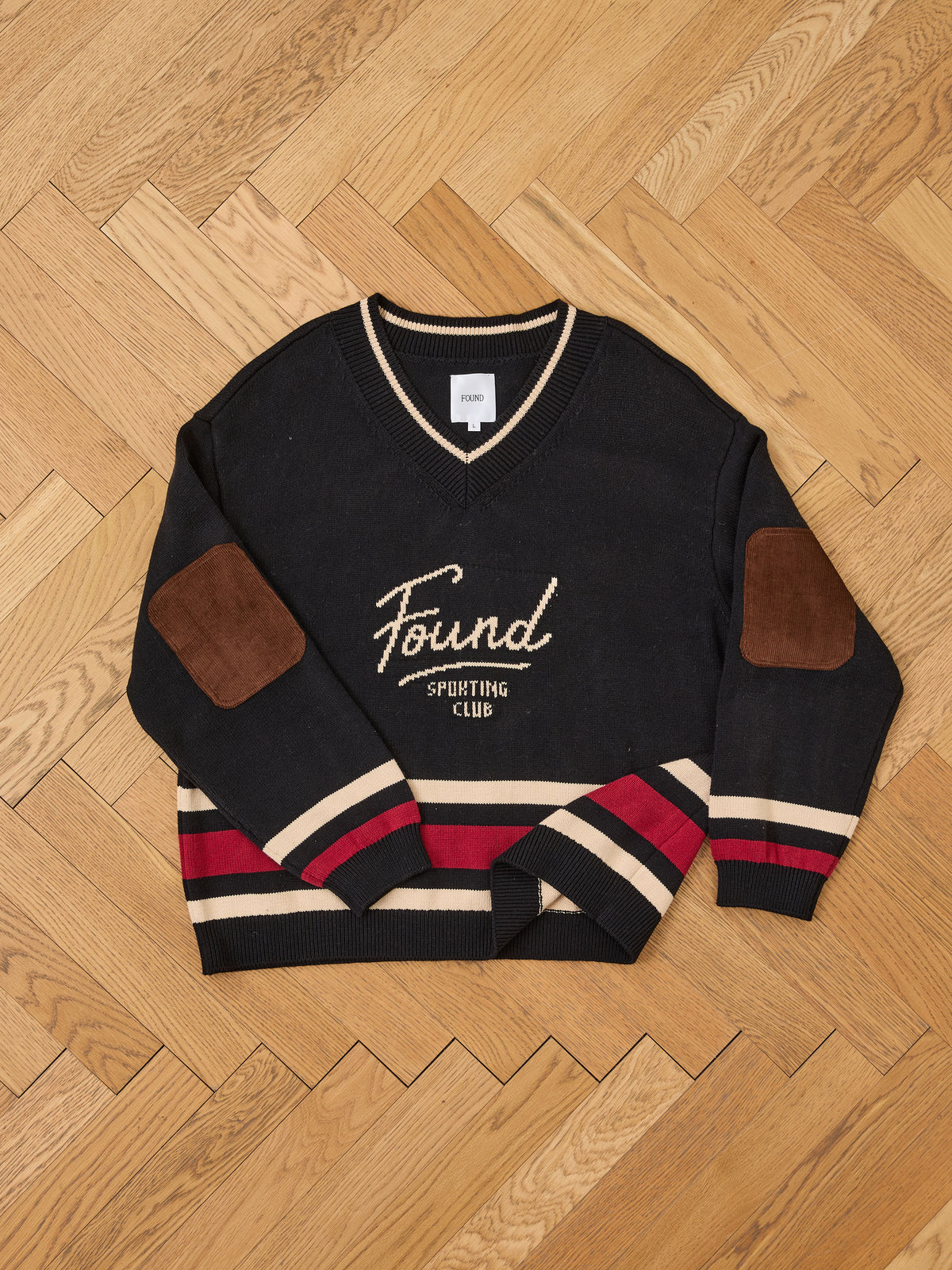 The Sport Club Varsity Sweater by FOUND is a black unisex relaxed-fit with “Found Sporting Club” text, red/beige stripes, and brown suede elbow patches.