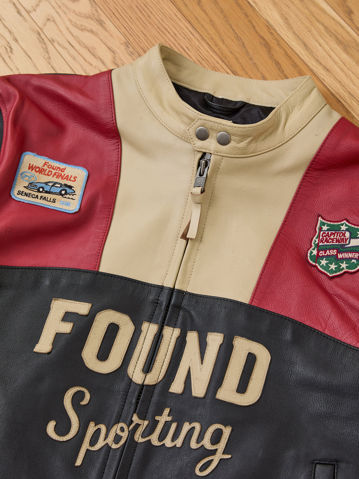 FOUND Sport Racer Jacket with front logo, two racing patches, and a tan, red, and black color block design on a wood floor.