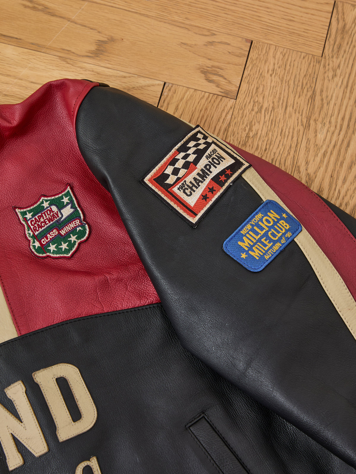The FOUND Sport Racer Jacket features red and black panels with three sewn patches, including a racing champion and “Million Mile Club,” shown on a wooden floor.