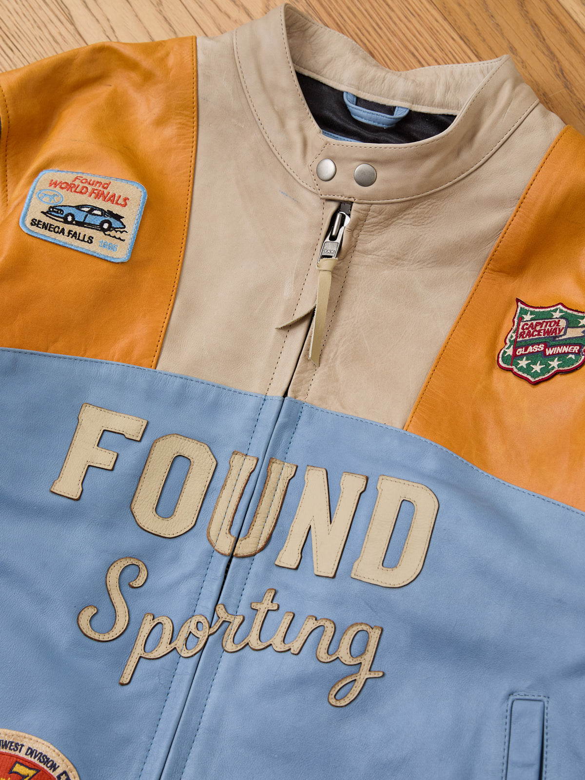 The FOUND Sport Racer Jacket, featuring blue, beige, and orange panels with “FOUND Sporting” embroidery and racing patches, lies flat on a wooden surface.