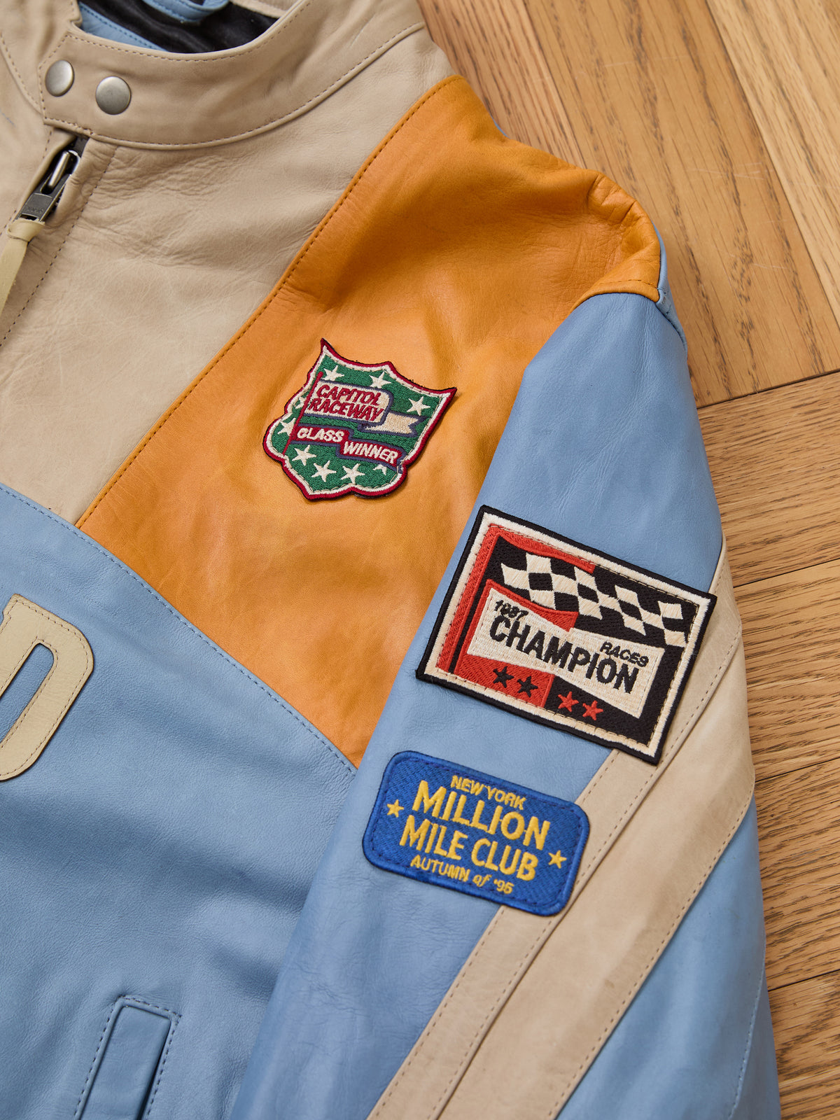 The FOUND Sport Racer Jacket in multicolored leather features racing-inspired patches like “Champion” and “Class Winner,” displayed on a wooden surface.