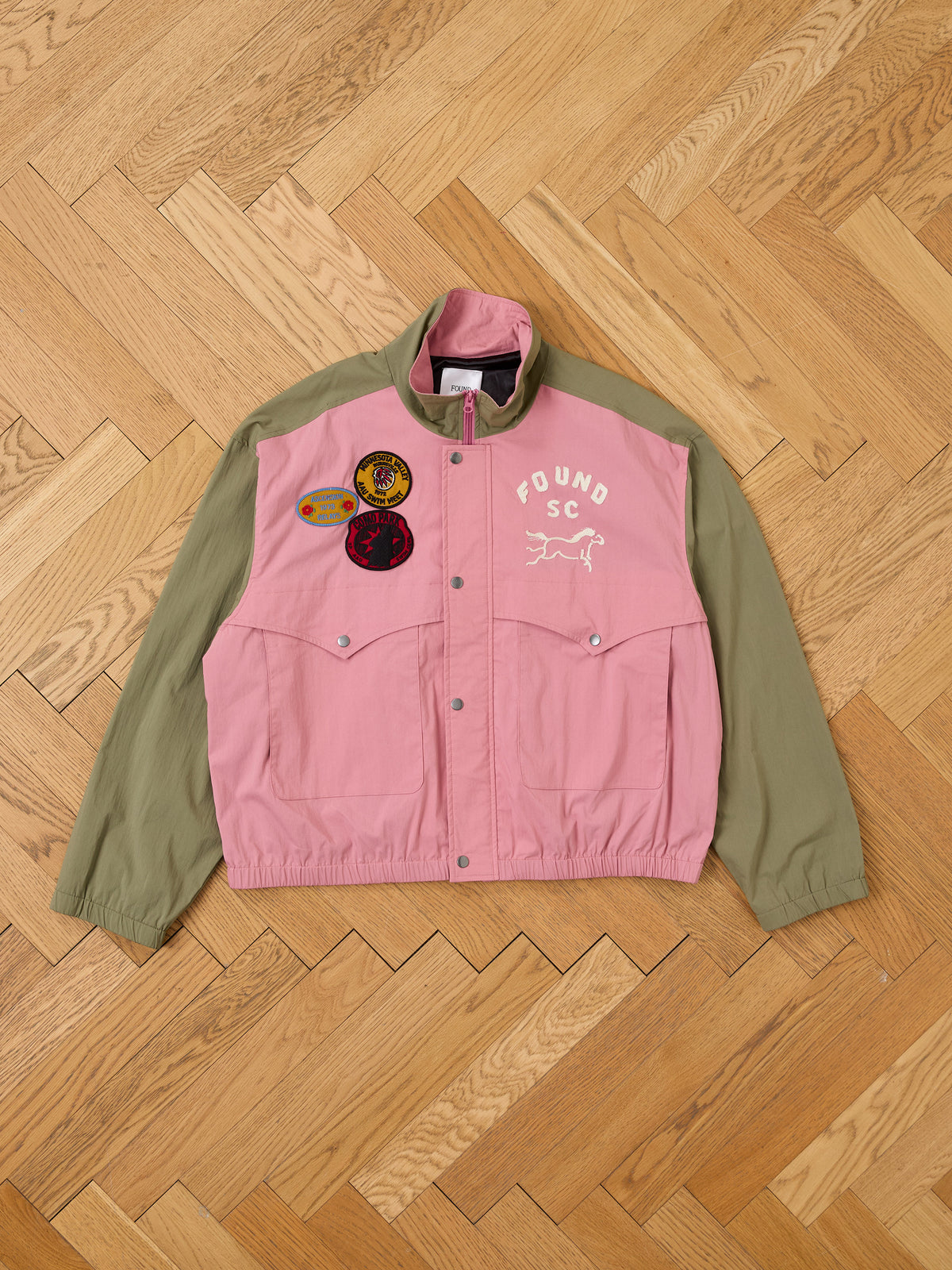 The Sport Windbreaker Jacket by FOUND, in pink and olive green with three patches, “FOUND SC” text, and a dog graphic, is displayed flat on a wooden herringbone floor.