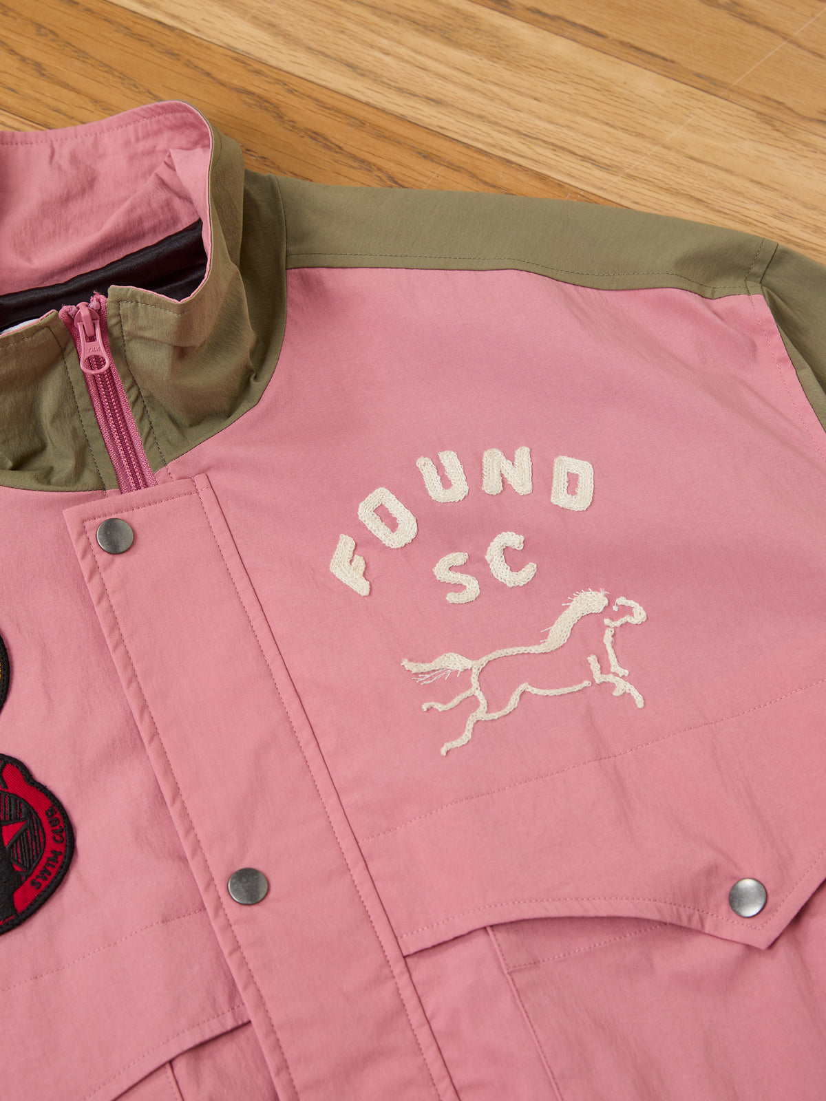 A pink and olive Sport Windbreaker Jacket by FOUND with “FOUND SC” and a horse embroidered on the chest, displayed on a wooden floor.