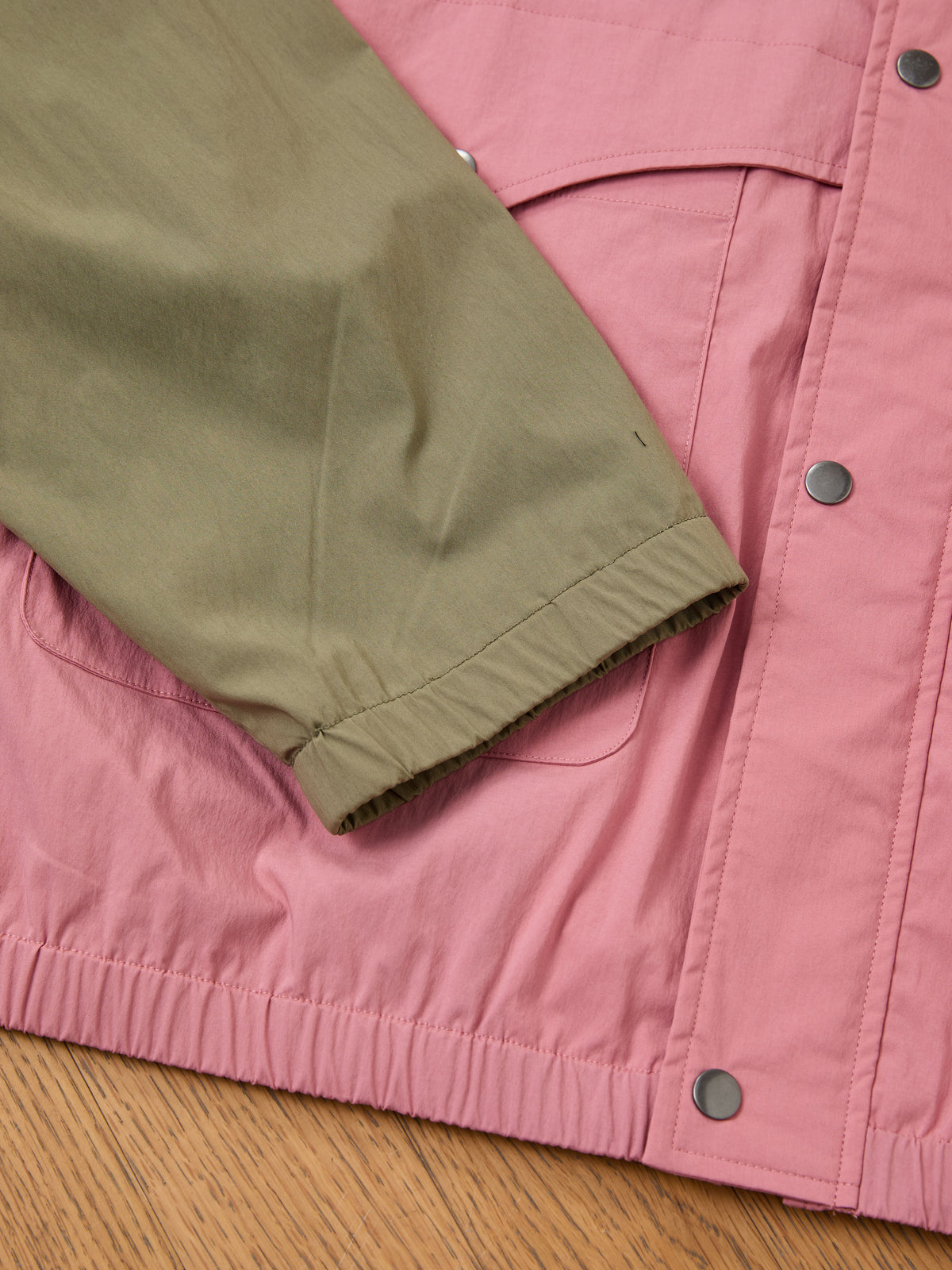 FOUND Sport Windbreaker Jacket in pink with silver snap buttons and olive green sleeve, displayed flat on a wooden surface.