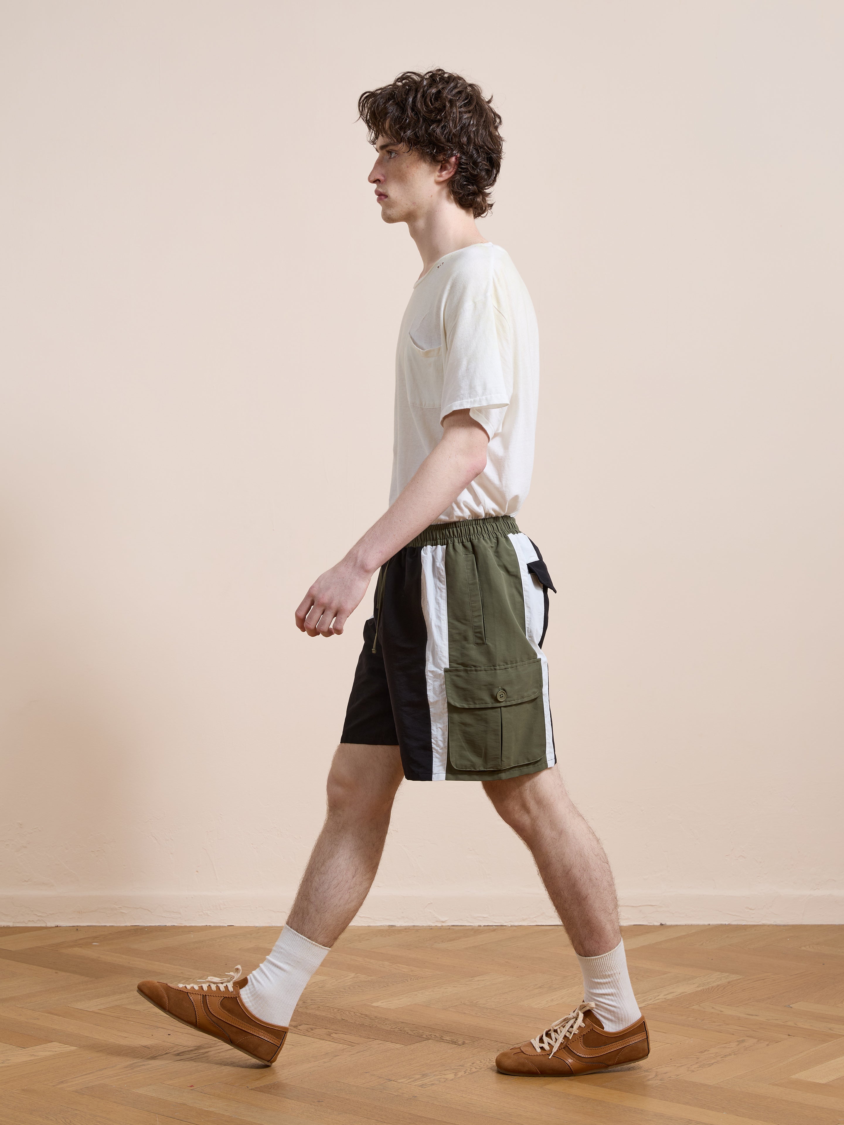 A man wearing a white t-shirt, Found Sporting Club Track Shorts in vintage green and white, white socks, and brown shoes walks indoors on a wooden floor with a plain beige wall in the background.
