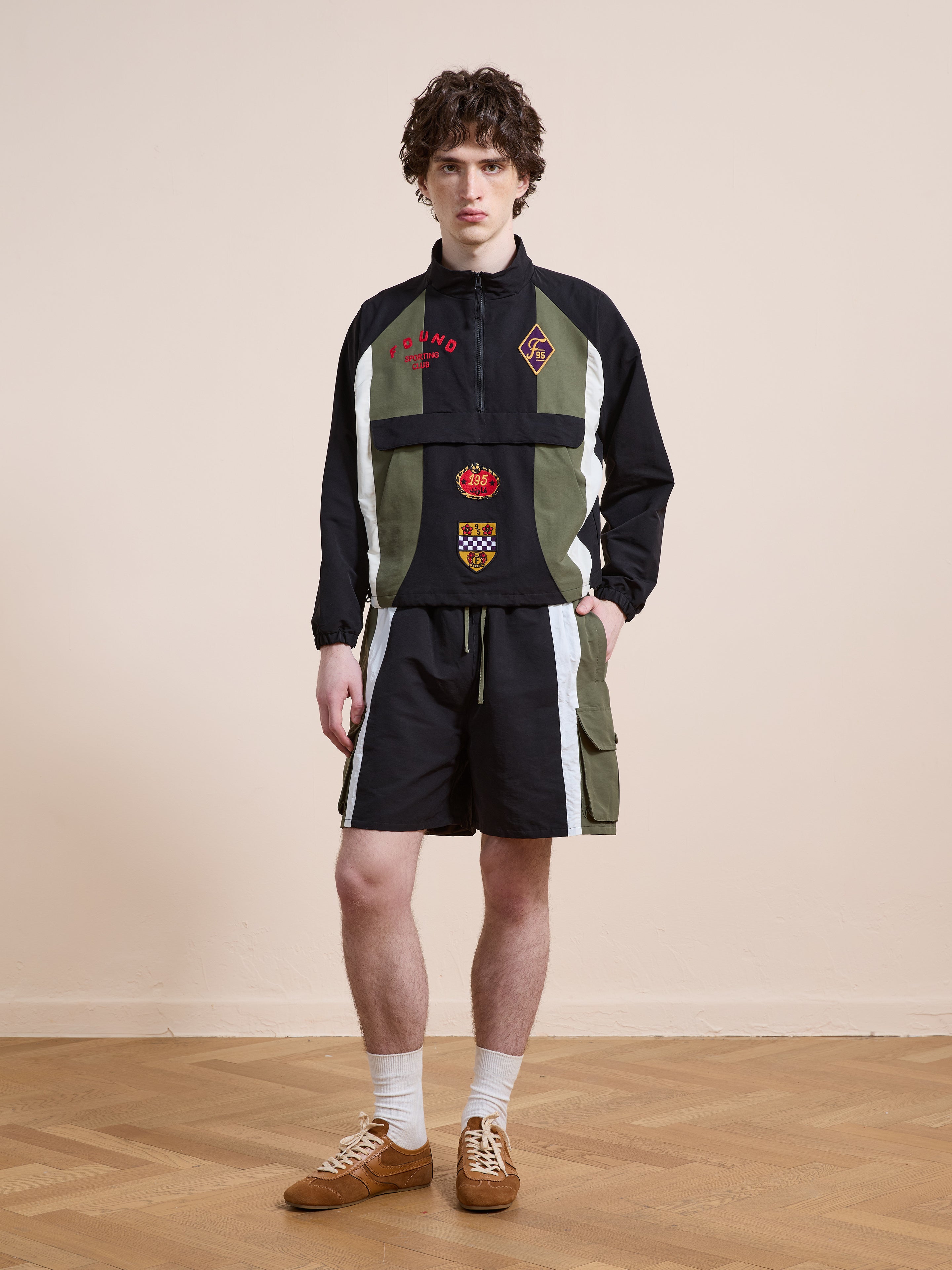 A young man stands on a parquet floor, wearing a vintage black and green long-sleeve top with emblems, Found Sporting Club Track Shorts in black with white stripes, white socks, and brown shoes.