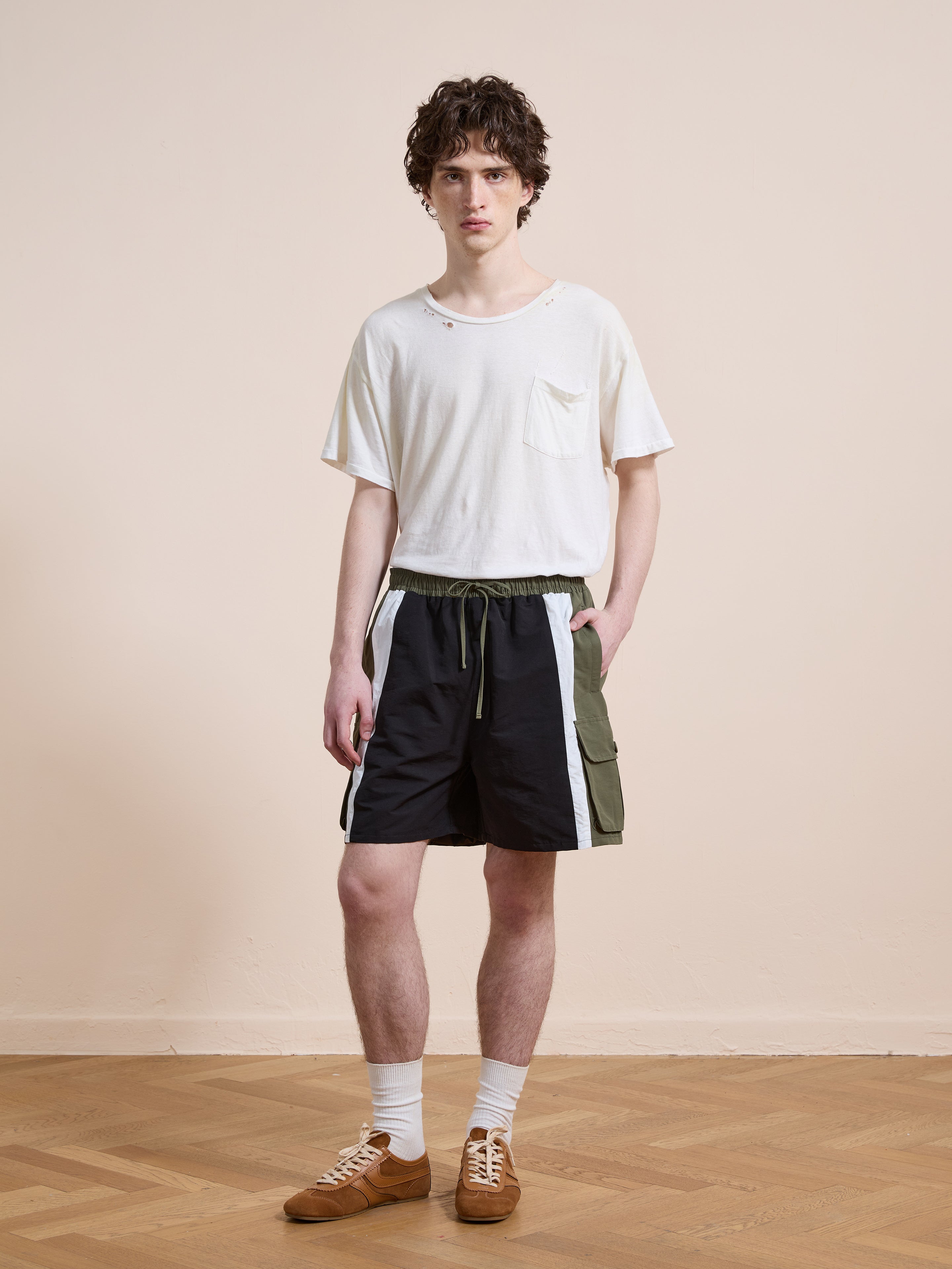 A person stands indoors wearing Found’s Sporting Club Track Shorts, styled with a white t-shirt, white socks, and brown sneakers. The unisex look radiates timeless sporty style against a beige wall and wooden floor.