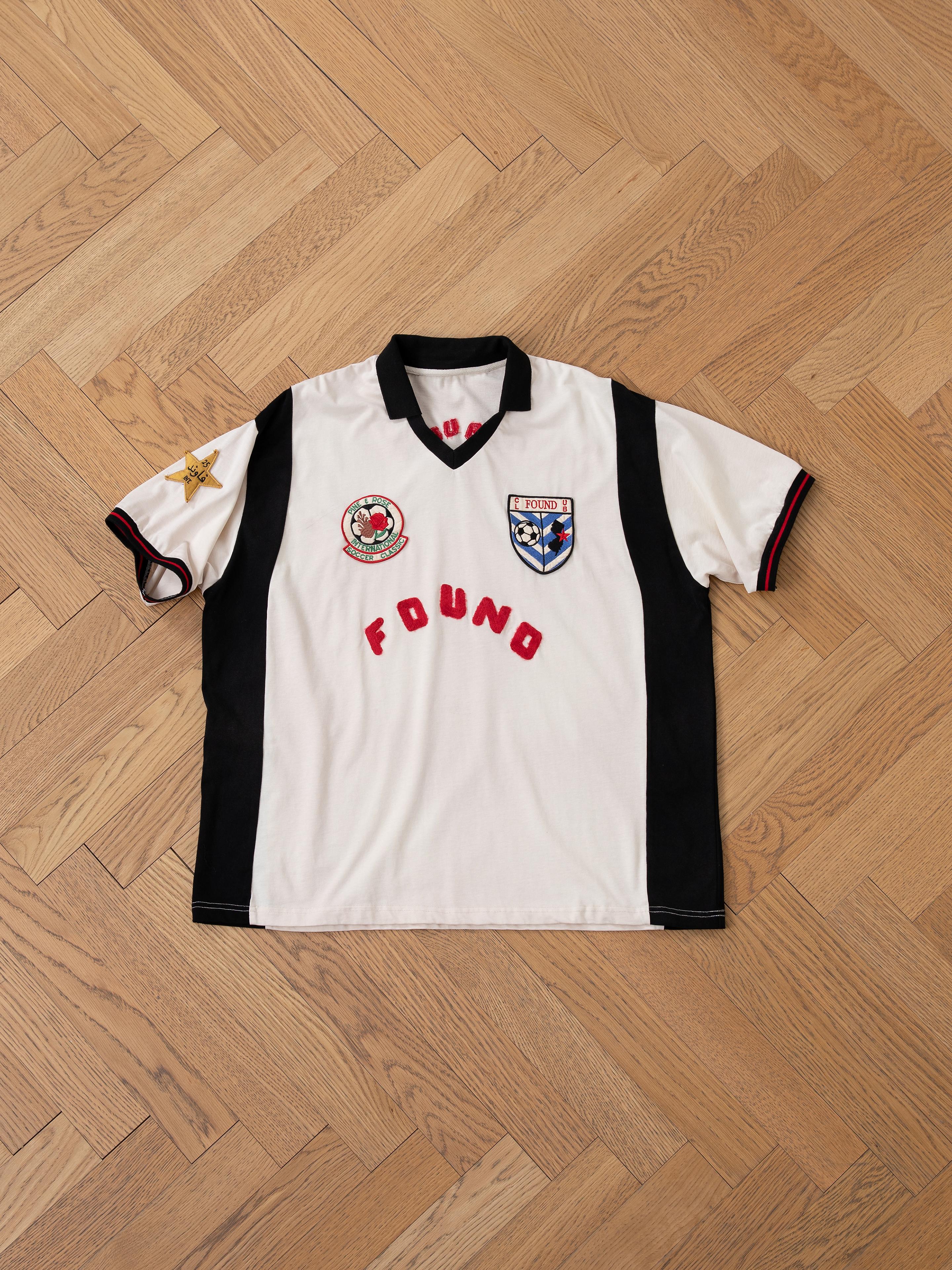 The FOUND Union Soccer Jersey, vintage-inspired and featuring a star patch on the left sleeve and two embroidered badges on the chest, is displayed on a wooden floor in white with black short sleeves.