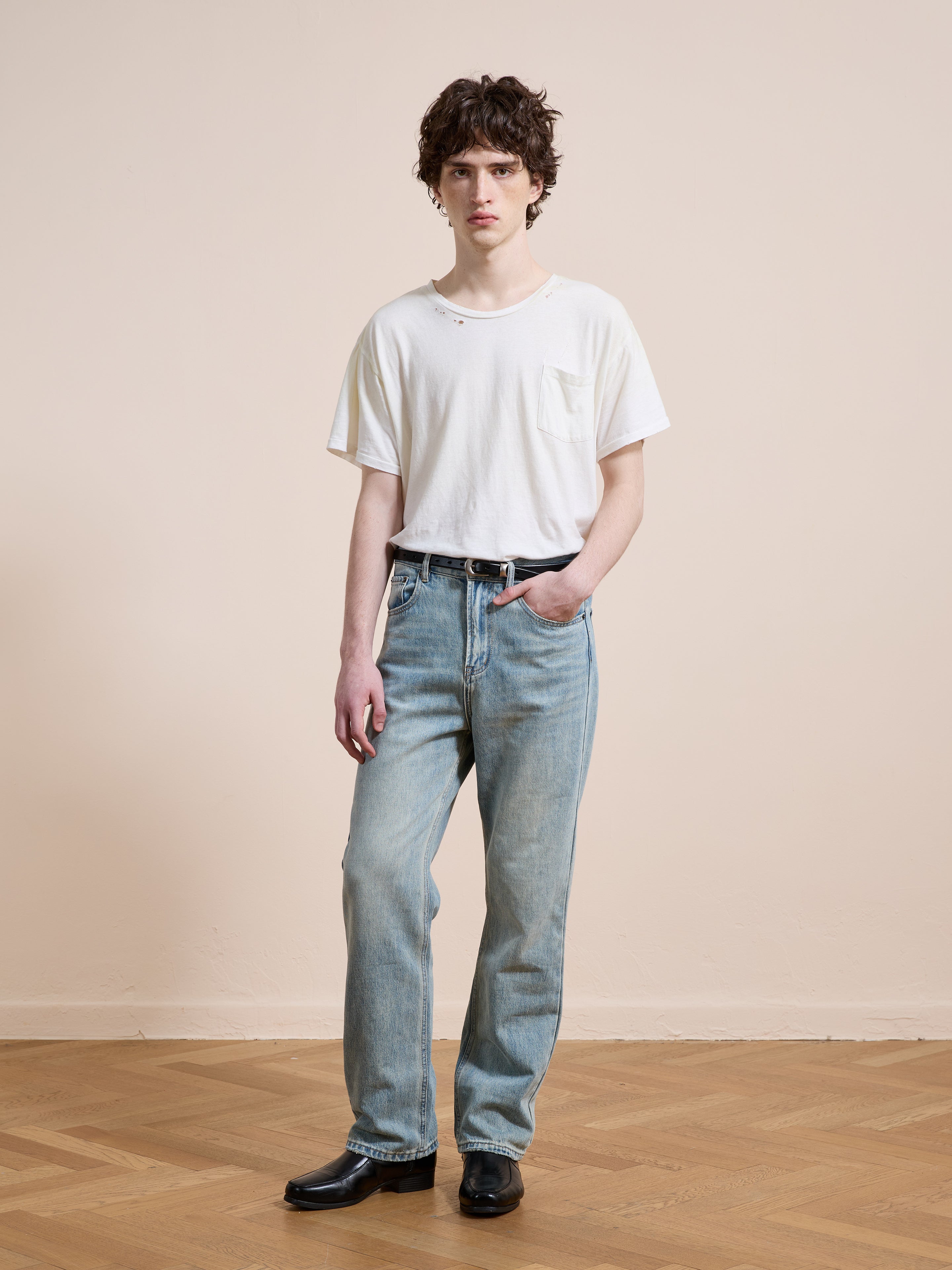 A person stands indoors on a wooden floor, wearing a white t-shirt, black belt, black shoes, and Found Straight Leg Jeans, facing the camera against a plain beige wall.