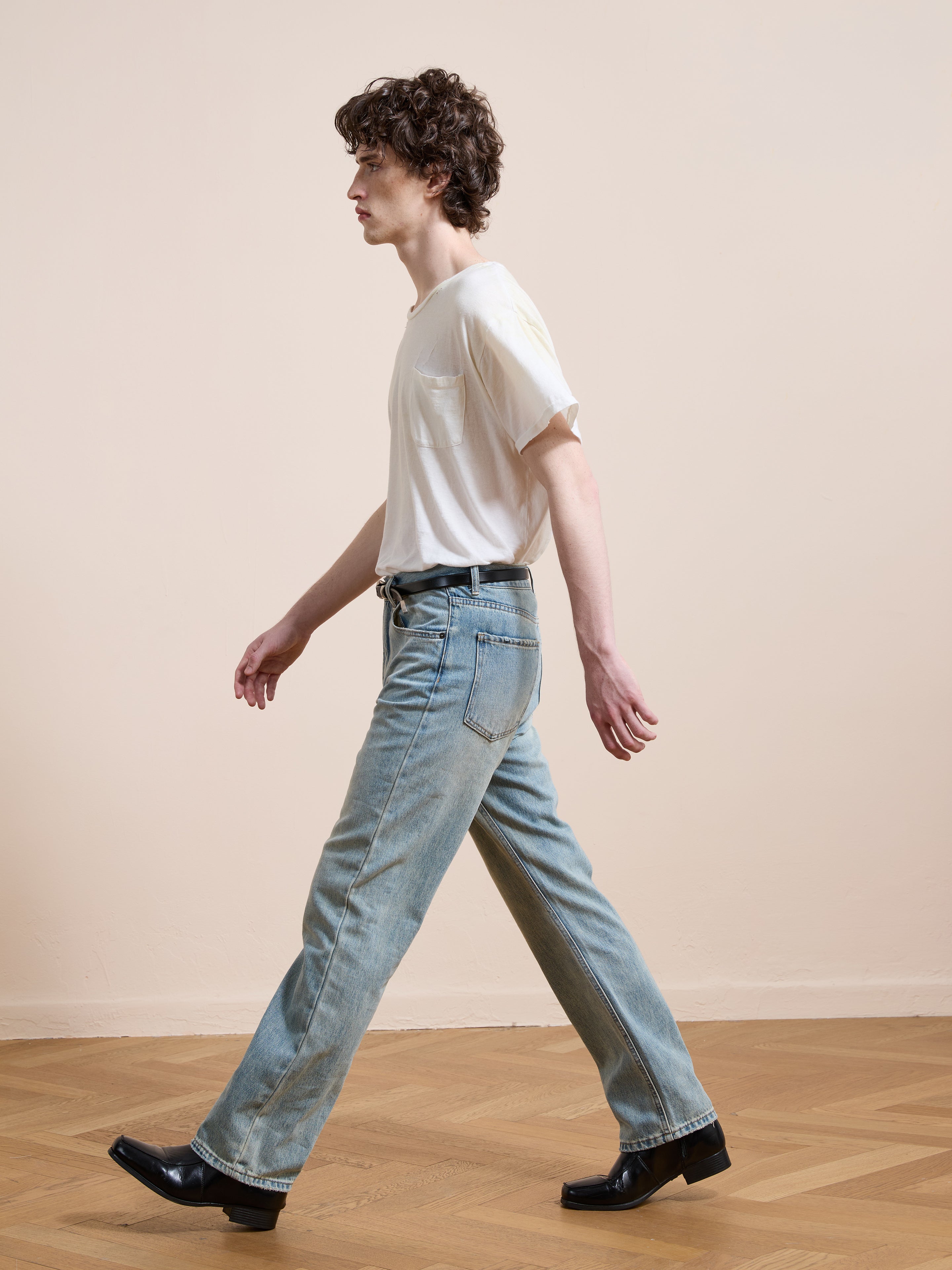 A person with curly hair walks indoors on a wooden floor, wearing a white t-shirt, black shoes, and Found Straight Leg Jeans.