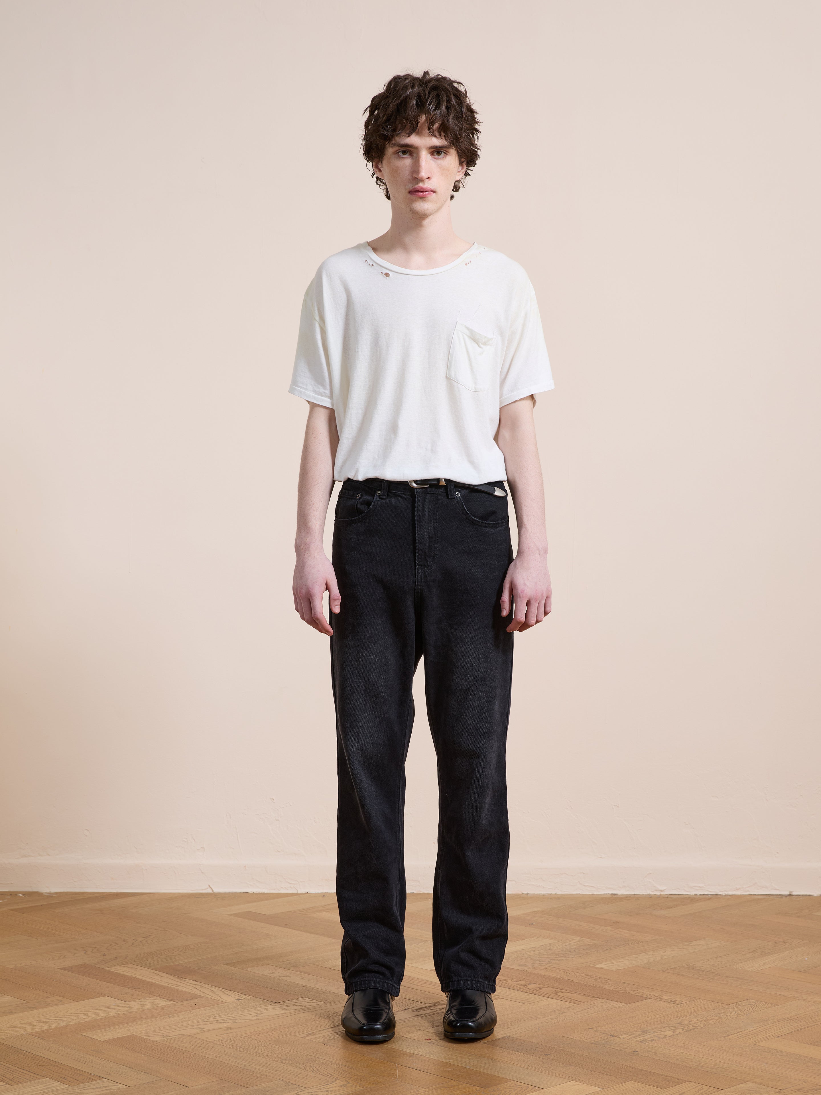 A person stands indoors on a wooden floor, wearing a white T-shirt with small holes, Found Straight Leg Jeans in black cotton denim, and black shoes, against a plain beige wall.