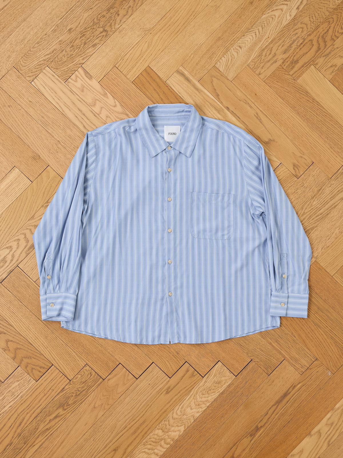 A Stripe Pocket Oxford Shirt by FOUND in light blue and white is laid flat, highlighting its unisex relaxed fit and classic chest pocket.