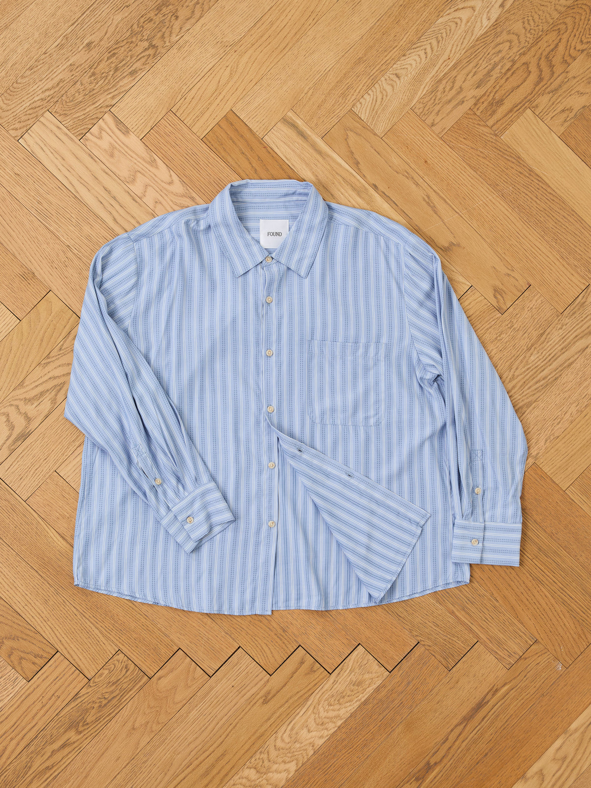 FOUND Stripe Pocket Oxford Shirt in light blue and white, unisex relaxed fit with front pocket, laid flat on a wooden herringbone floor.