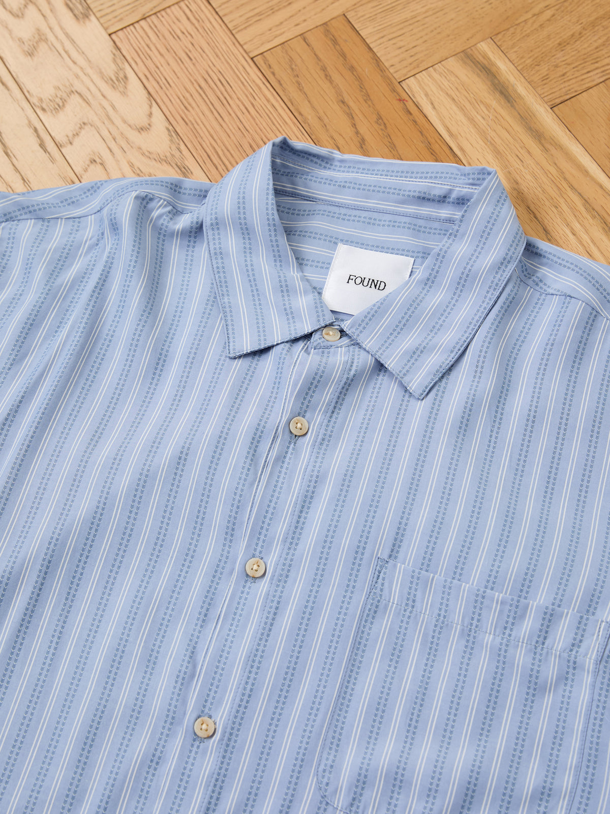 The Stripe Pocket Oxford Shirt by FOUND is a light blue unisex relaxed fit button-up with white stripes and a pocket, shown flat on a wooden floor.