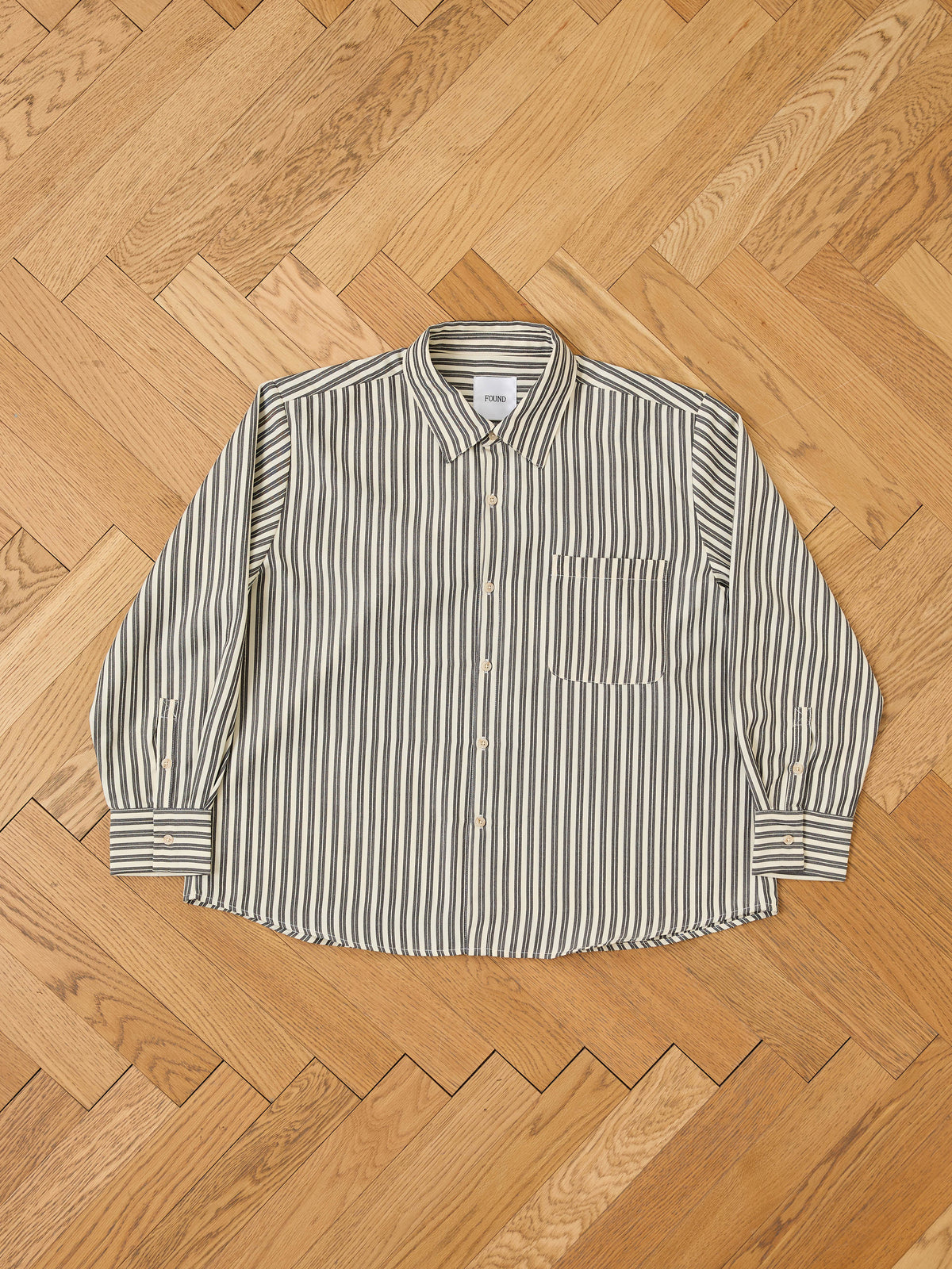 The FOUND Stripe Pocket Oxford Shirt with vertical black and white stripes and a unisex relaxed fit is laid flat on a herringbone-patterned wooden floor.