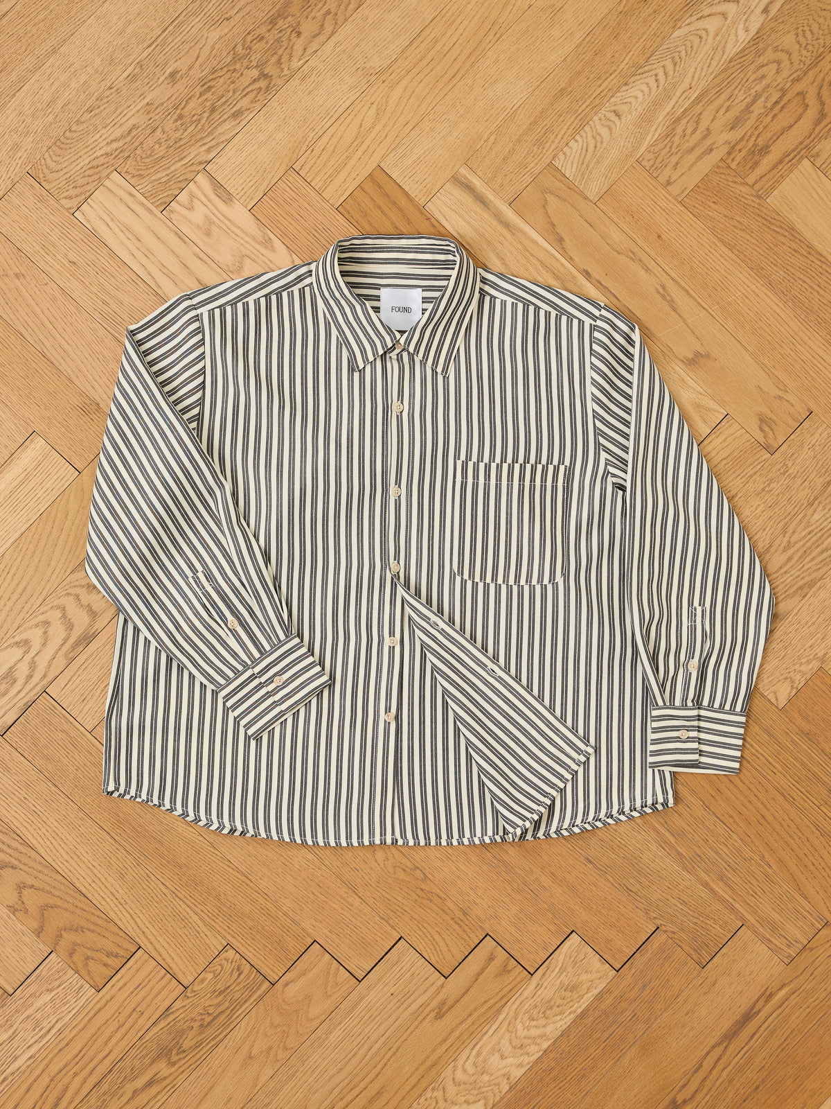 The FOUND Stripe Pocket Oxford Shirt features vertical stripes and a unisex relaxed fit, shown laid flat on a light wood herringbone floor.