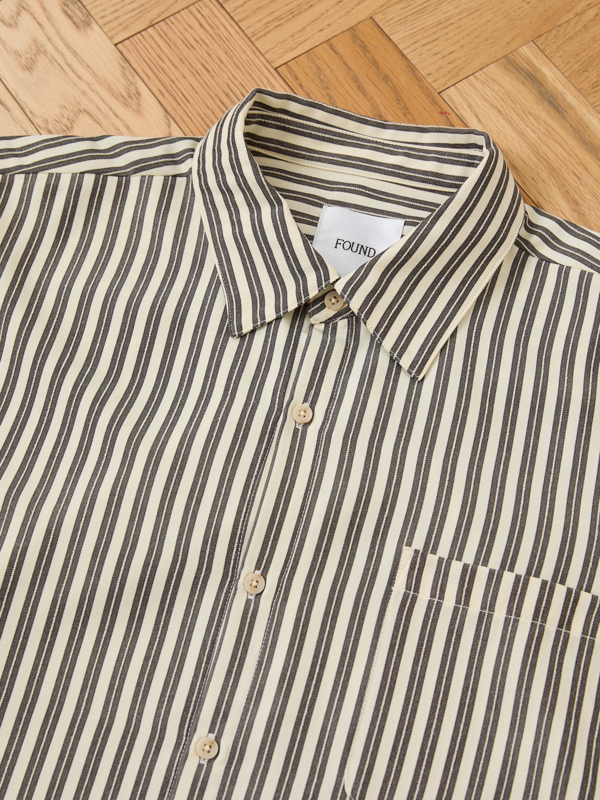 A FOUND Stripe Pocket Oxford Shirt in beige with black vertical stripes is laid flat, showing its collar, buttons, and unisex relaxed fit.