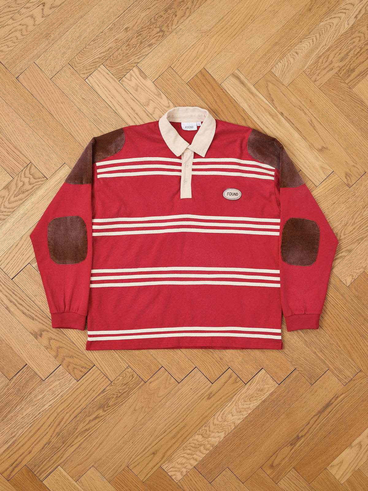 The FOUND Striped Corduroy Rugby Polo is a unisex, relaxed-fit shirt in red and white stripes with brown suede elbow patches, a beige collar, and a small oval patch reading “YOUNG.” Crafted from midweight cotton, it is shown laid flat on a wooden floor.