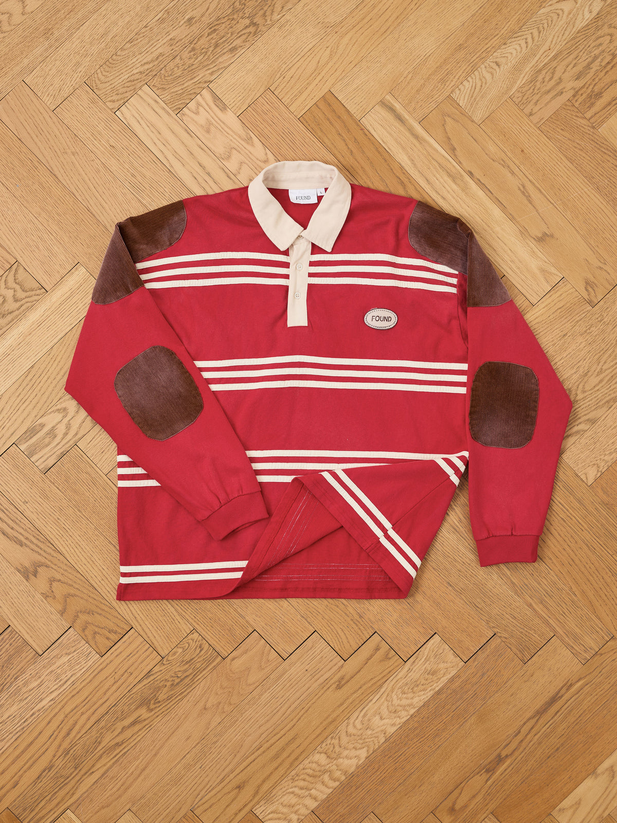 FOUND’s Striped Corduroy Rugby Polo in red features a beige collar, horizontal white stripes, and brown corduroy elbow patches. Crafted from midweight cotton with a relaxed unisex fit, this rugby shirt is adorned with a small “FOUND” patch on the chest and is shown laid flat on a wooden floor.