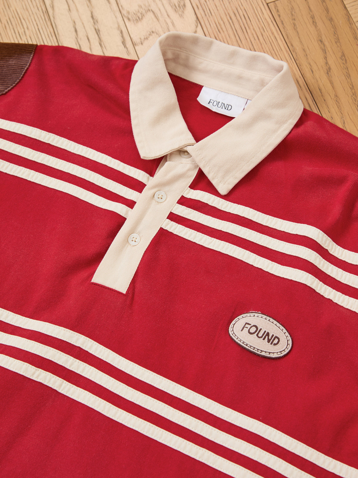 The FOUND Striped Corduroy Rugby Polo features a red corduroy body with a beige collar and stripes, three-button placket, dual “FOUND” labels, and is crafted from midweight cotton in a unisex relaxed fit—shown here on a wooden floor.