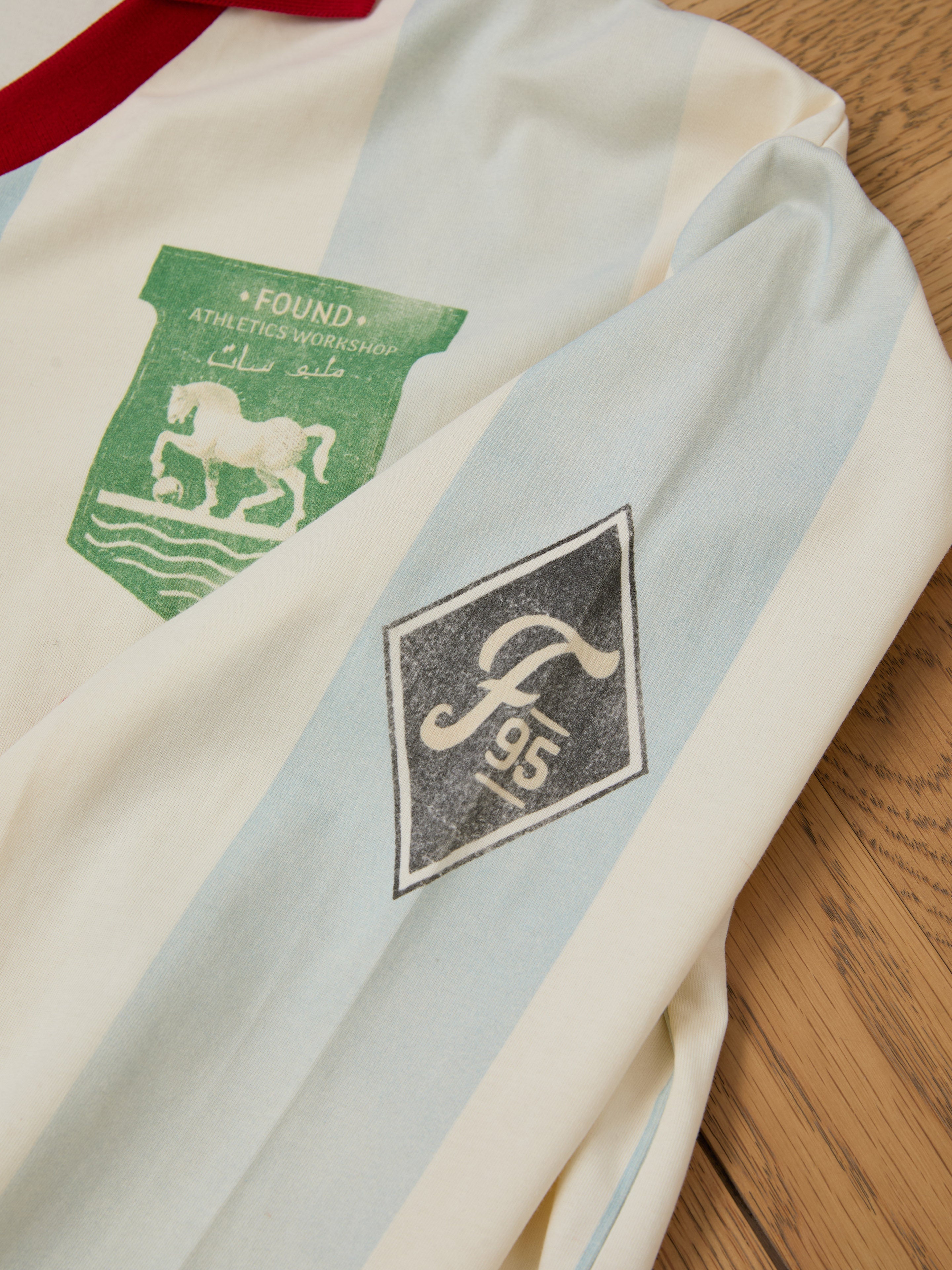 The Striped Sport LS Jersey by FOUND lies on a wooden surface, featuring a vintage sportswear vibe with a green horse emblem and an F95 badge on the sleeve.