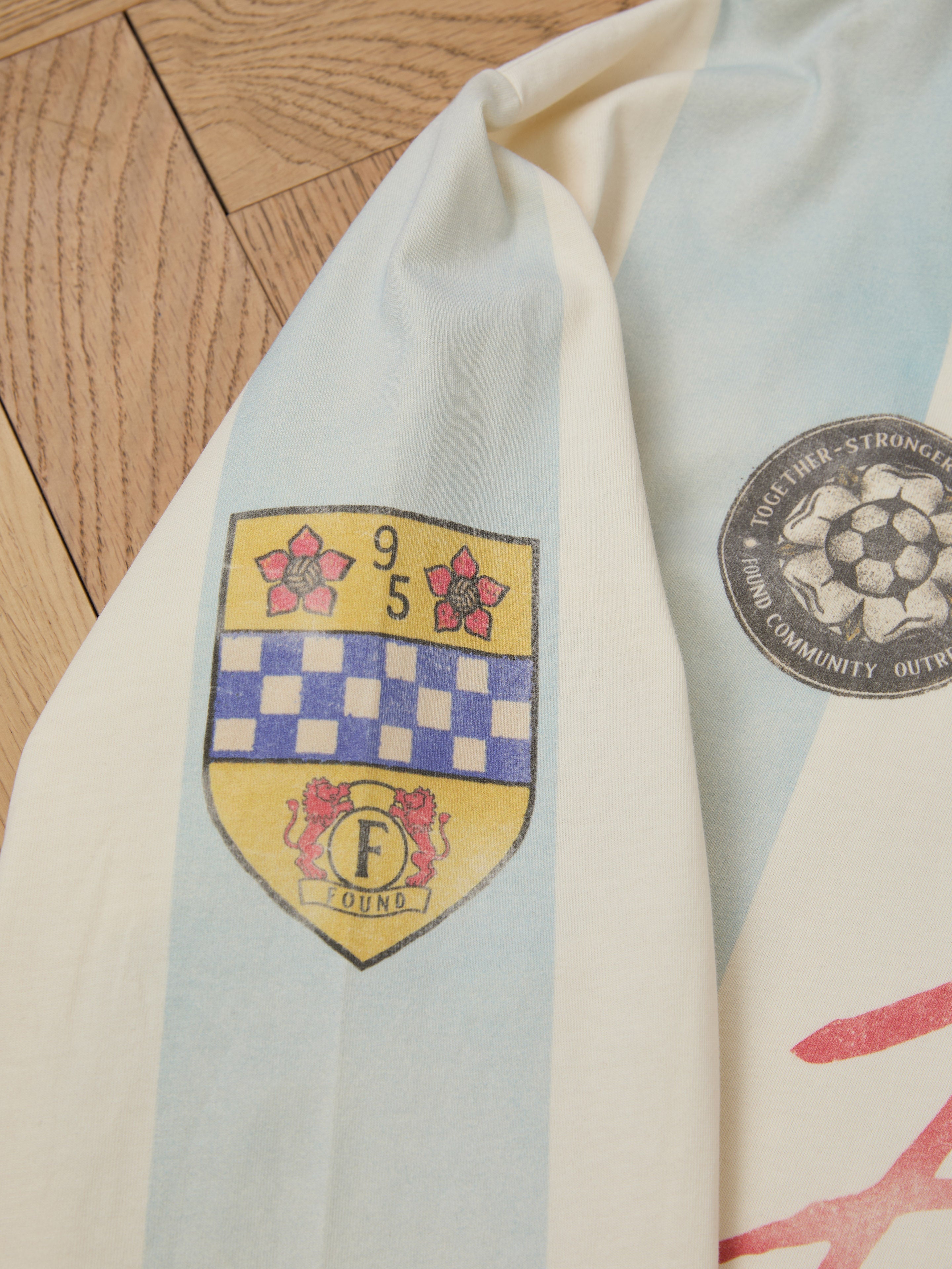 A Striped Sport LS Jersey by FOUND, featuring a vintage logo with two red flowers, a blue checkered pattern, the number 95, the letter "F," and a round emblem with text and a soccer ball symbol, lies on a wooden surface.