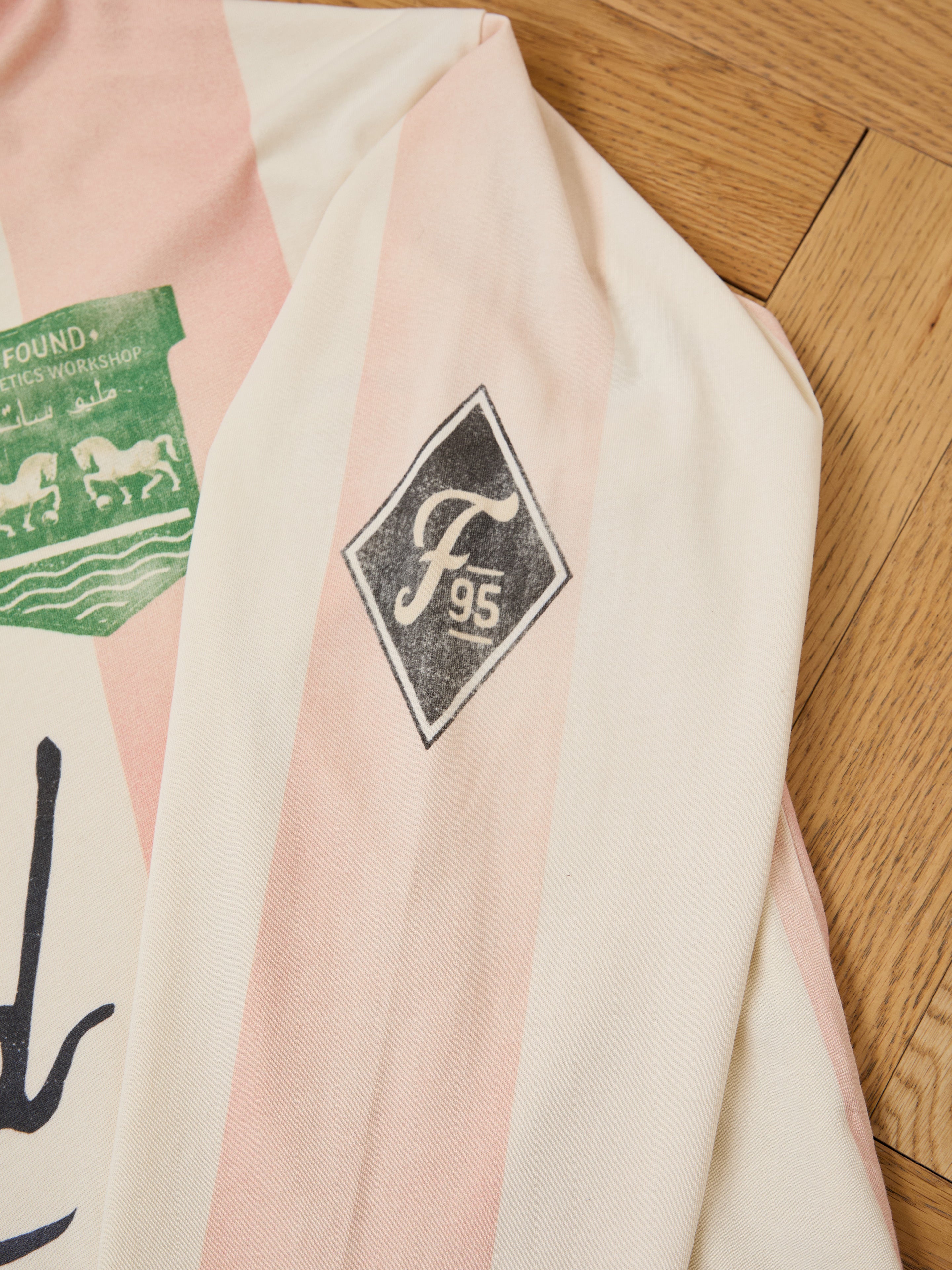 A close-up of the FOUND Striped Sport LS Jersey with pink and white vertical stripes, a black diamond-shaped "F95" logo on the sleeve, and part of a green chest graphic, laid on a wooden floor.