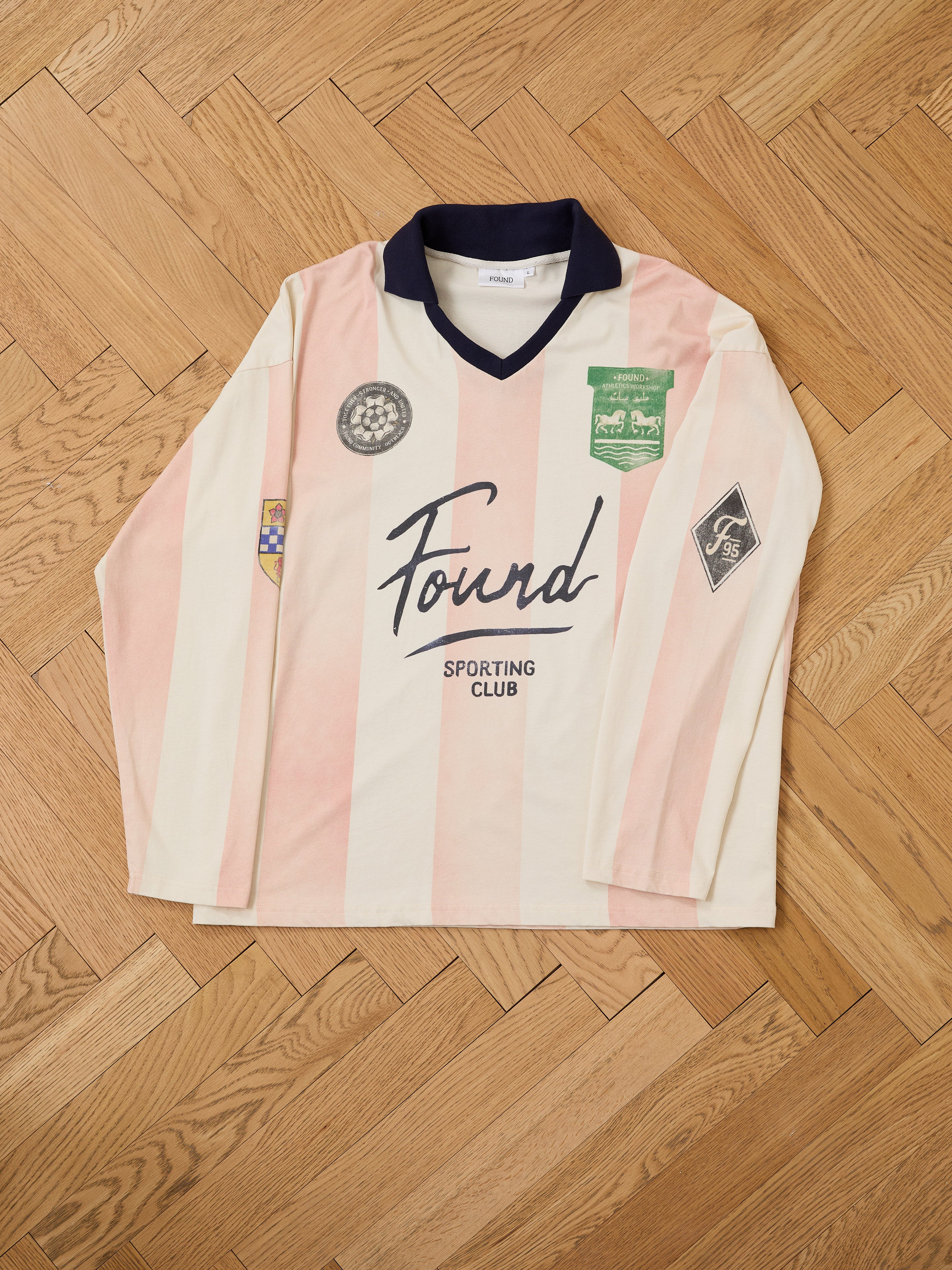 The FOUND Striped Sport LS Jersey is a unisex, relaxed-fit long-sleeve top in cream and pink stripes with patches and "Found Sporting Club" on the front, shown on a wooden floor in a modern football-inspired style.